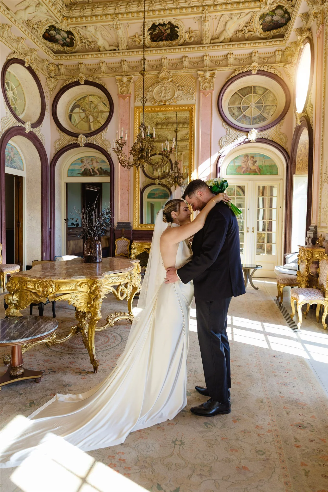 A bride and groom share a romantic moment in an ornate, historic room with intricate ceiling moldings and large windows. The bride is in a white wedding dress with a long train, and the groom is in a black suit. They are embracing and smiling at each