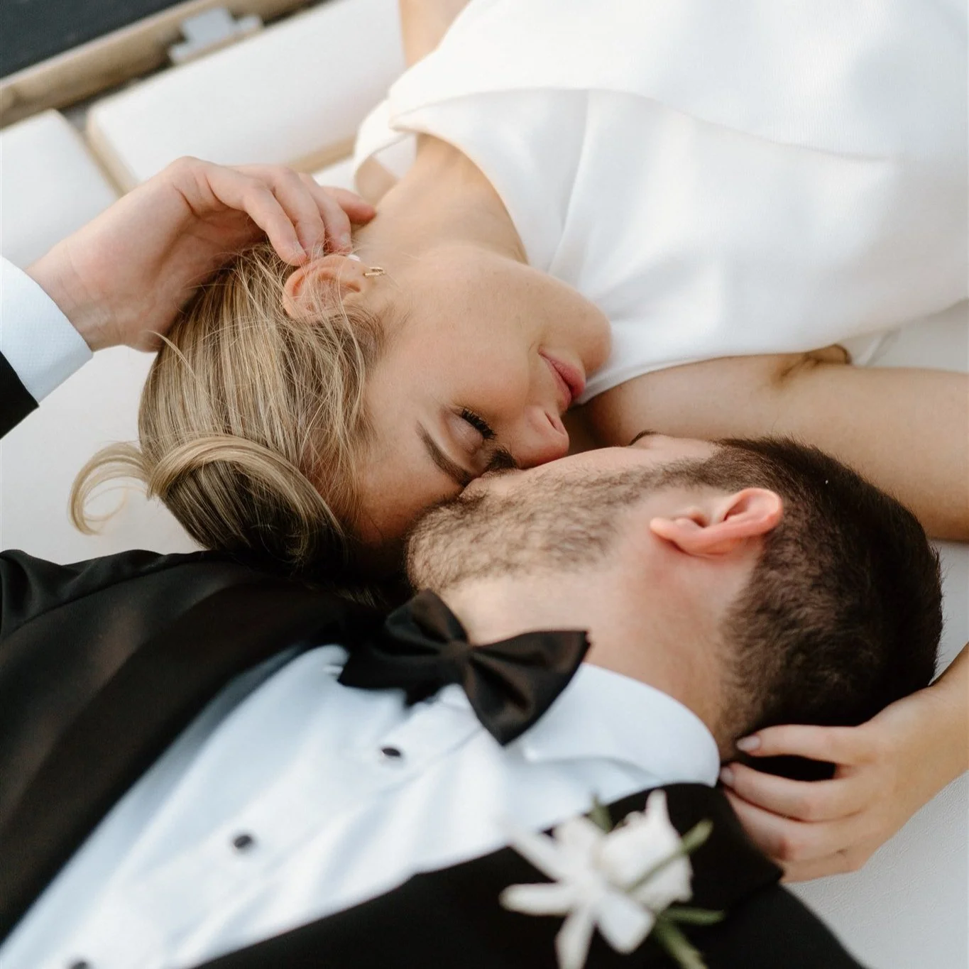 A couple is lying down and embracing each other. The woman has blonde hair and is wearing a white dress, with her eyes closed and touching the man's face. The man has dark hair and a beard, and is wearing a tuxedo with a bow tie. A person's hand is touching the woman's hair.