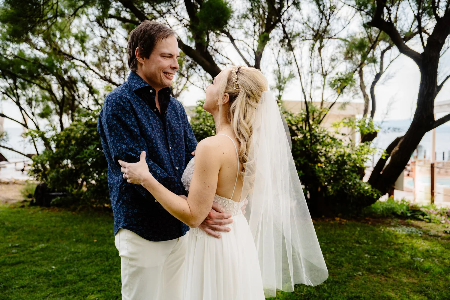 A happy couple, a man and a woman, standing outdoors on a grassy area with trees in the background, looking at each other and smiling. The woman is dressed in a wedding gown with a veil, and the man is wearing a dark blue patterned shirt and white pa