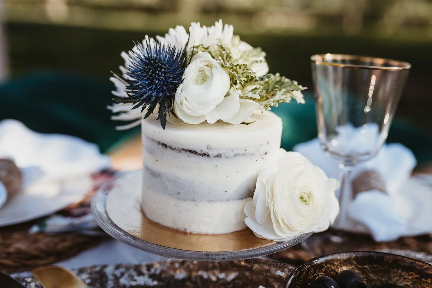 A small, white, two-layer naked cake decorated with white flowers and a dark blue thistle on top, set on a silver cake stand at an outdoor event.