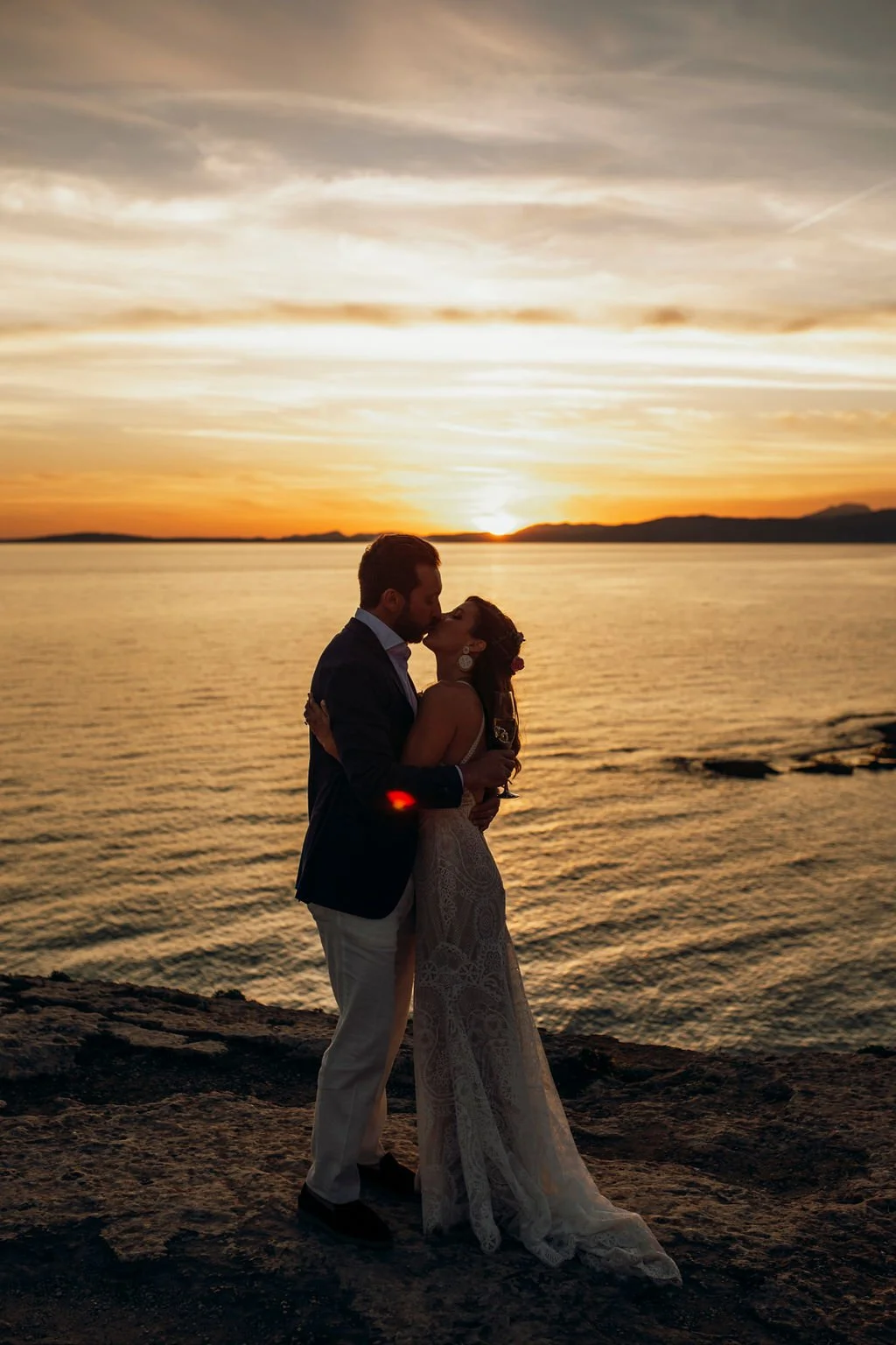 A couple kissing during sunset by the water. The man wears a suit and the woman wears a lace dress. The sky is filled with clouds and the sun is near the horizon, creating a romantic scene.