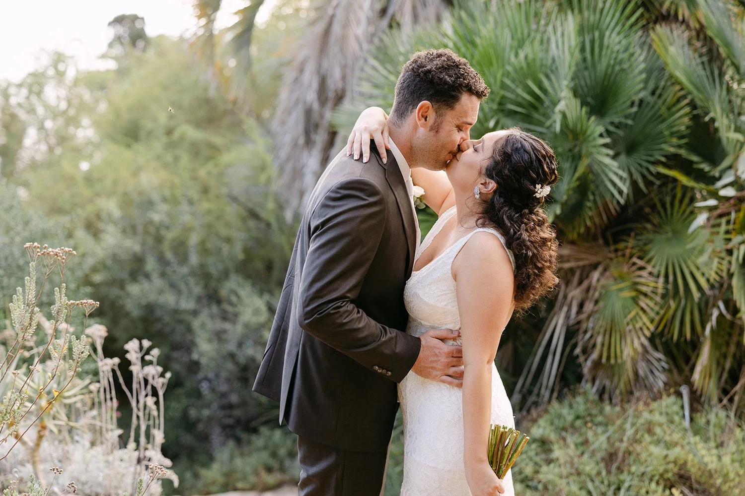 A newlywed couple sharing a kiss outdoors surrounded by greenery, with the bride holding a bouquet of flowers.