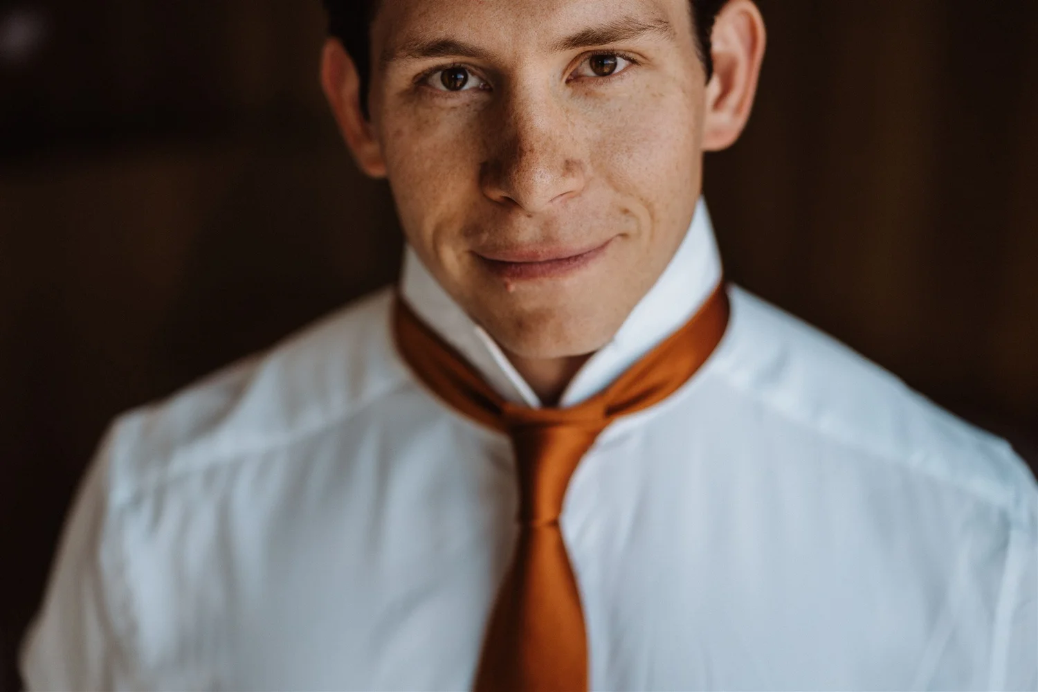 Close-up of a young man wearing a white shirt and orange tie, smiling slightly, with brown hair and freckles.