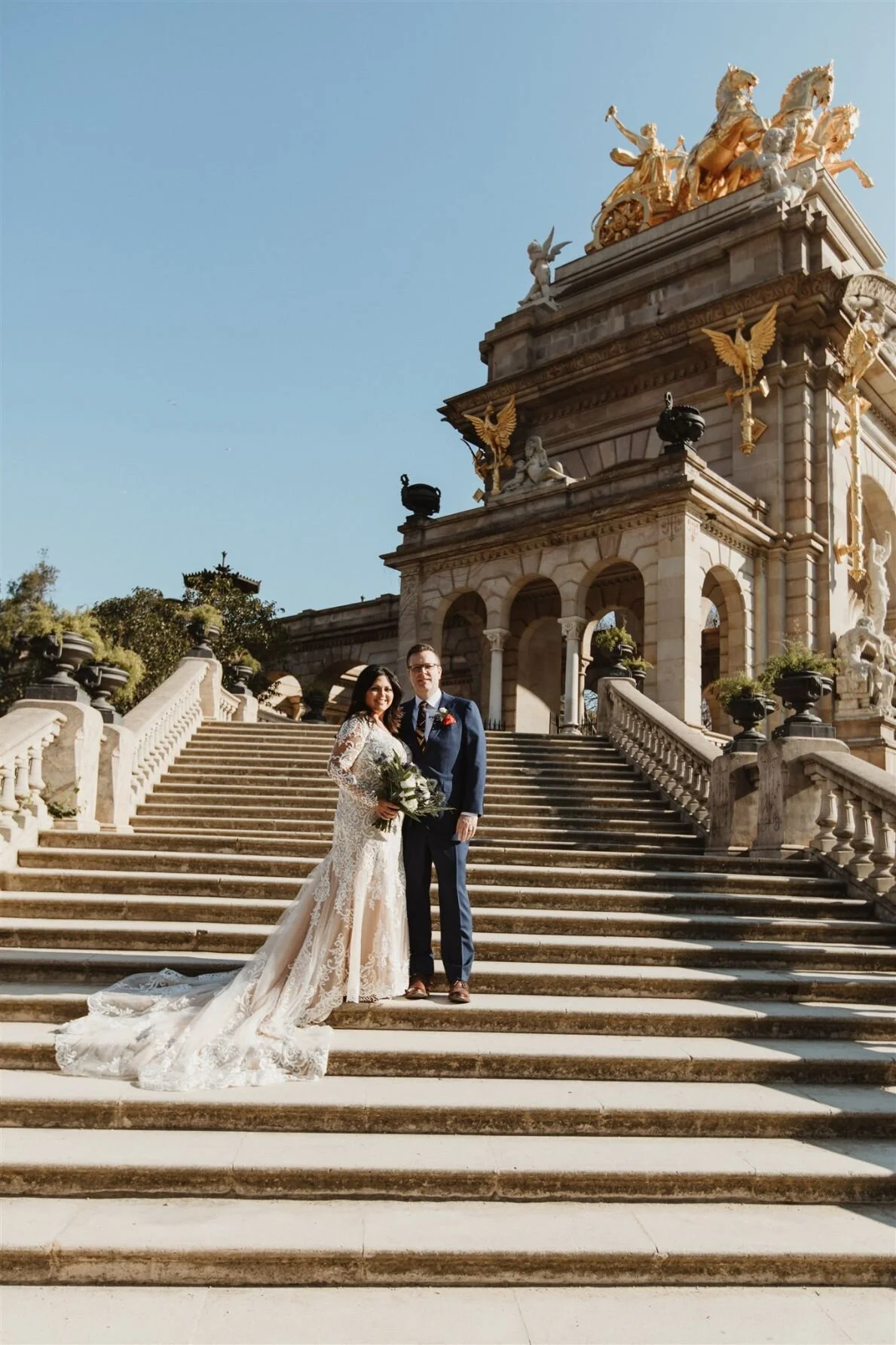 Bride and groom standing on steps in front of ornate historic building with sculptures and golden details, wedding attire, bride holding bouquet, sunny day.