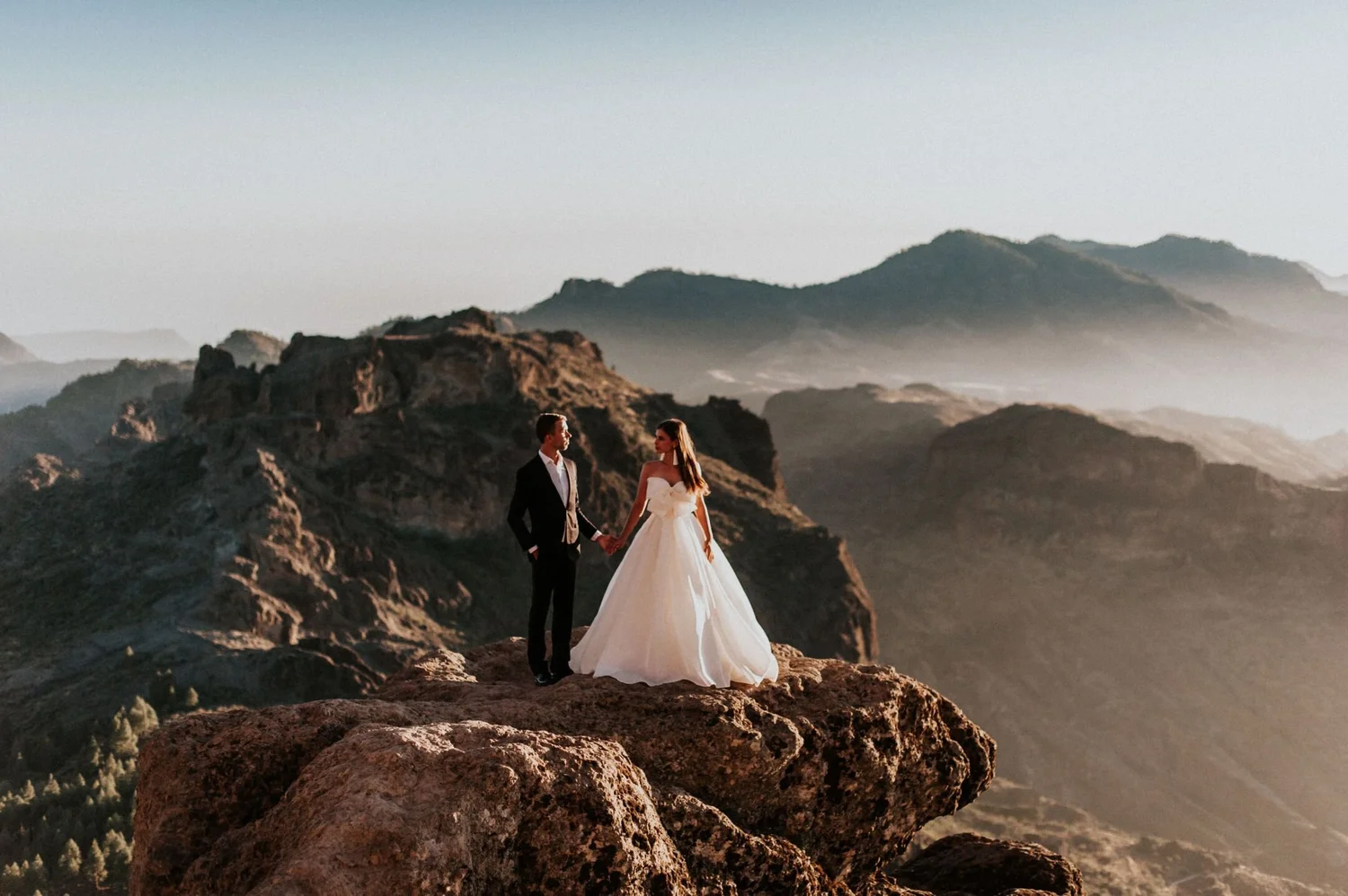 A bride and groom holding hands standing on rocky terrain with mountainous landscape in the background during sunset.