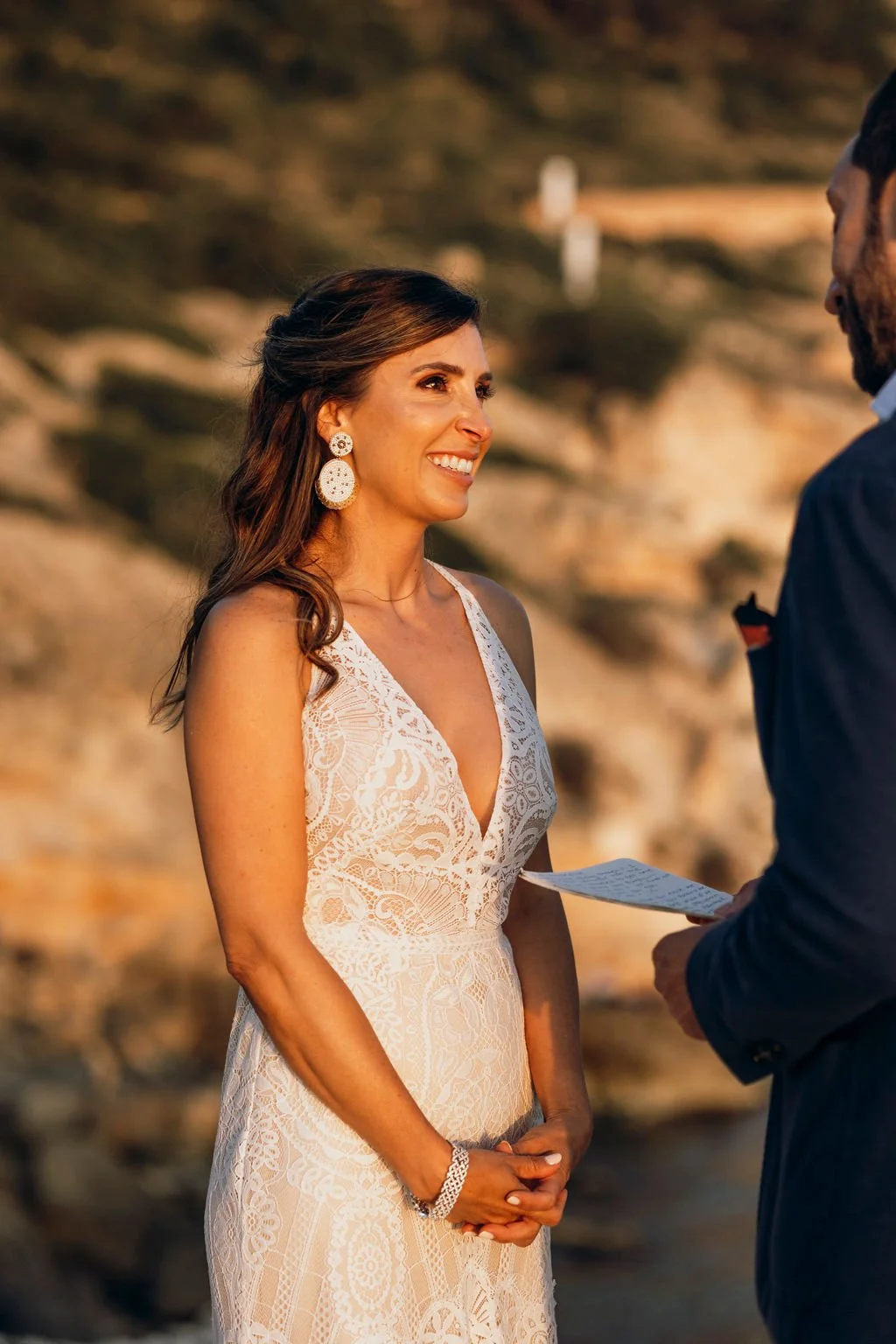 A woman in a white lace dress smiling at a man holding a piece of paper, outdoors during sunset with rocky terrain background.