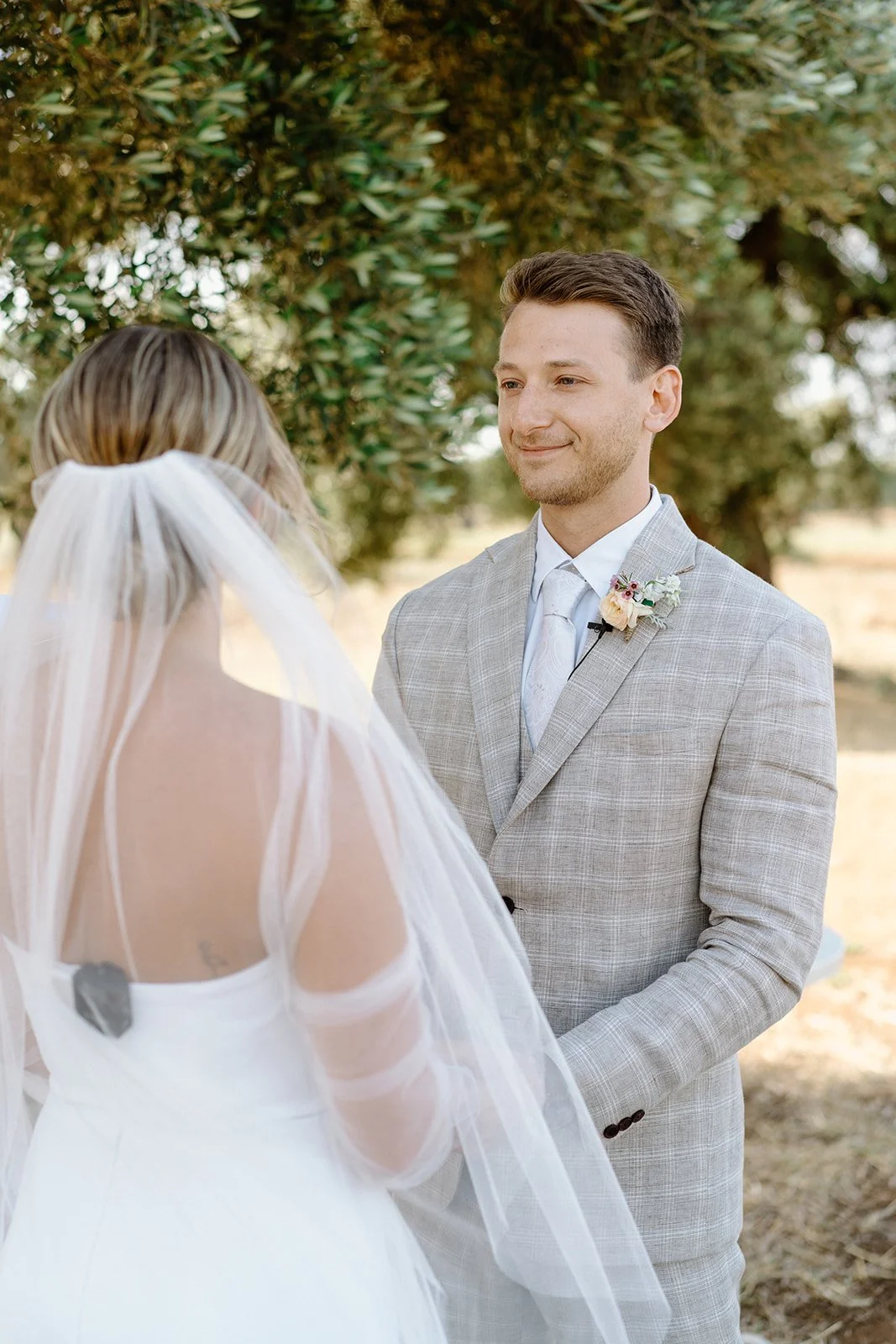 A bride and groom exchanging vows outdoors under a large tree, with the groom smiling and wearing a light gray suit with a boutonniere, and the bride wearing a white gown with a veil.