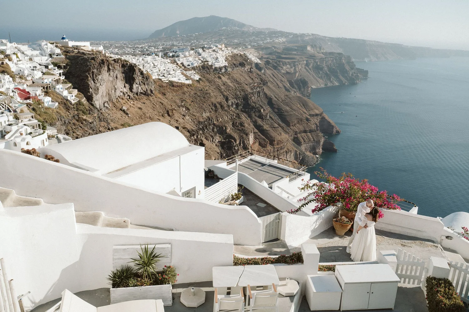 Two women dressed in white wedding gowns standing close together near a large potted plant with pink flowers on a white terrace overlooking the ocean and rugged cliffs in Santorini, Greece.