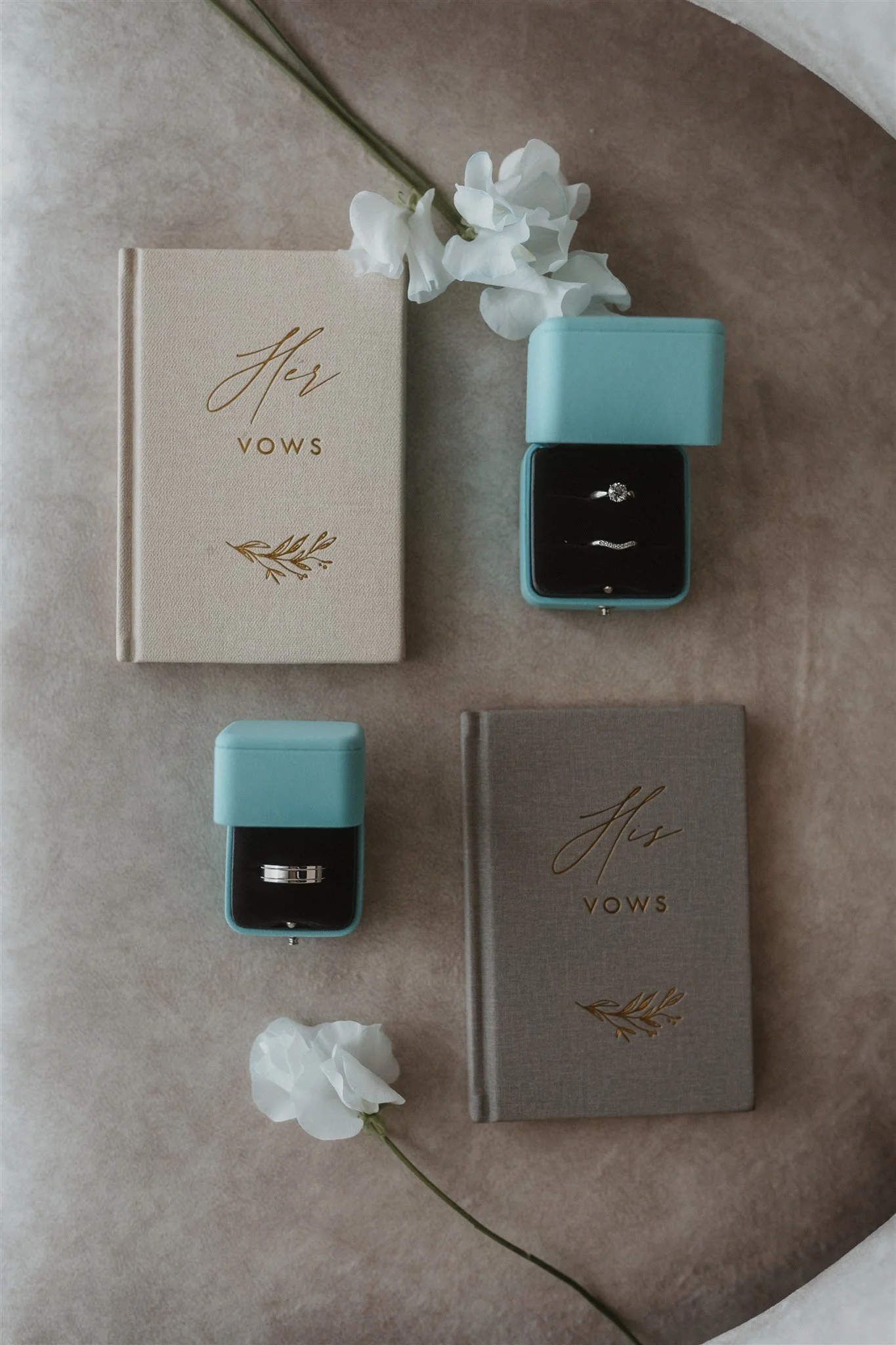 A wedding ring inside a turquoise box, a vows book, white flowers, tissue paper, and a beige background.