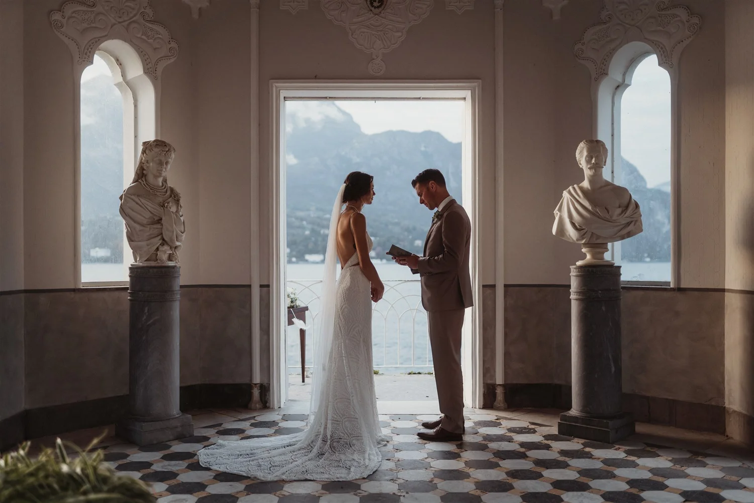 A bride and groom stand facing each other, holding hands, during their wedding ceremony indoors, with large windows showing mountains and water in the background. The setting features ornate decorations and classical statues.