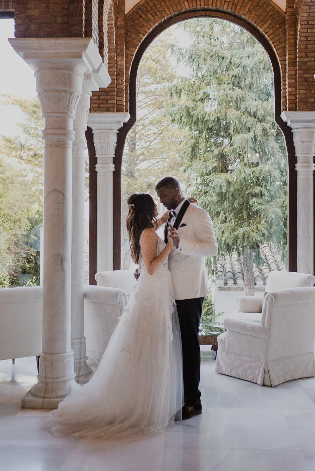 A bride and groom dancing together inside a grand room with large arched windows and white marble columns.