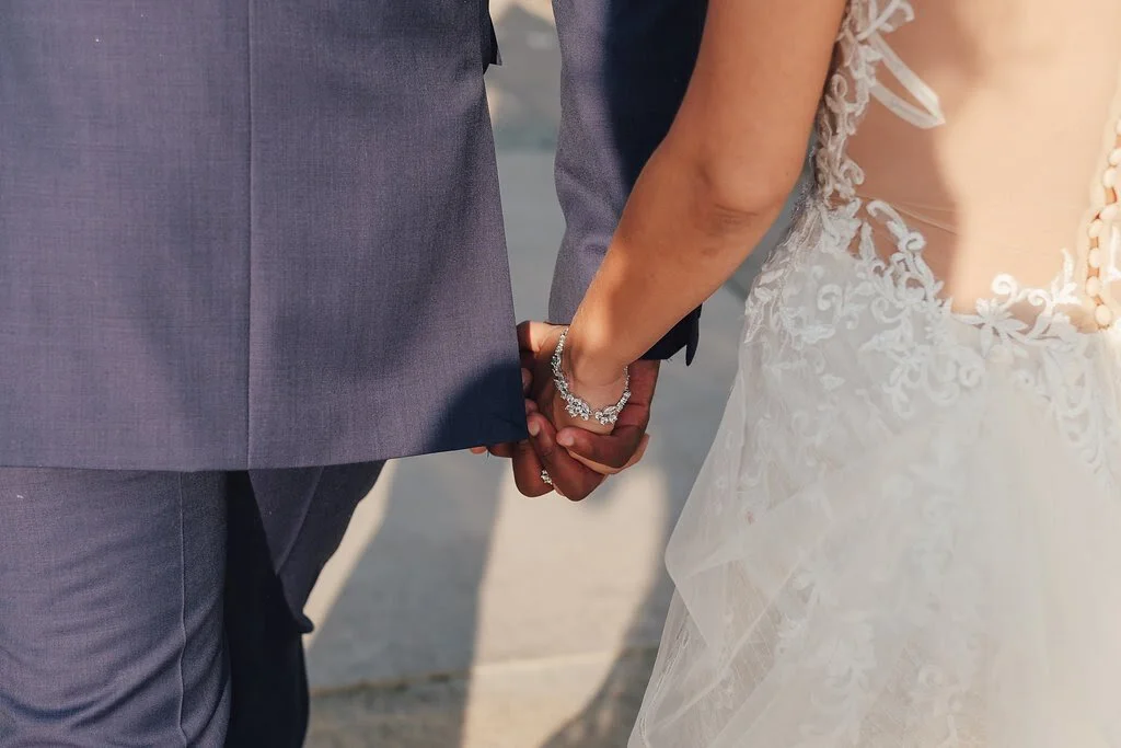 Close-up of a bride and groom holding hands, with the bride wearing a lace wedding dress and bracelet, and the groom in a gray suit.