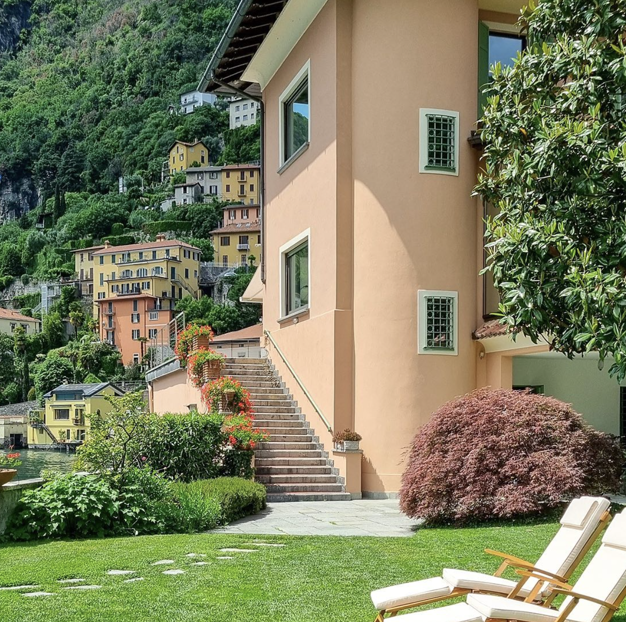 A view of a hillside residential area with colorful houses and lush greenery, as seen from a garden with a patio, stairs, and lounge chairs.