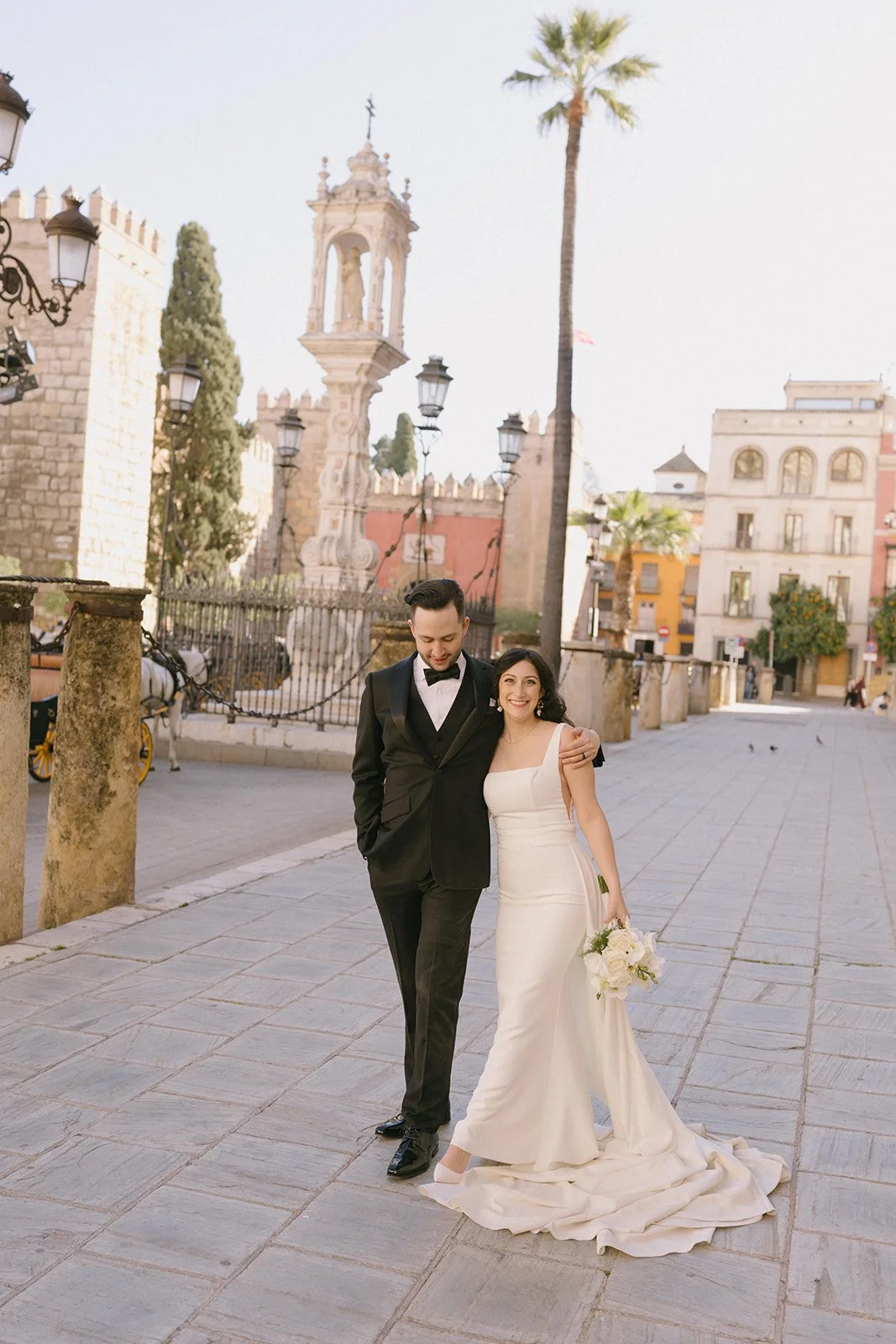 A newlywed couple walking in a historic city street with palm trees, old buildings, and a tall ornate fountain in the background. The groom in a black tuxedo and the bride in a white wedding gown holding a bouquet pose happily together.
