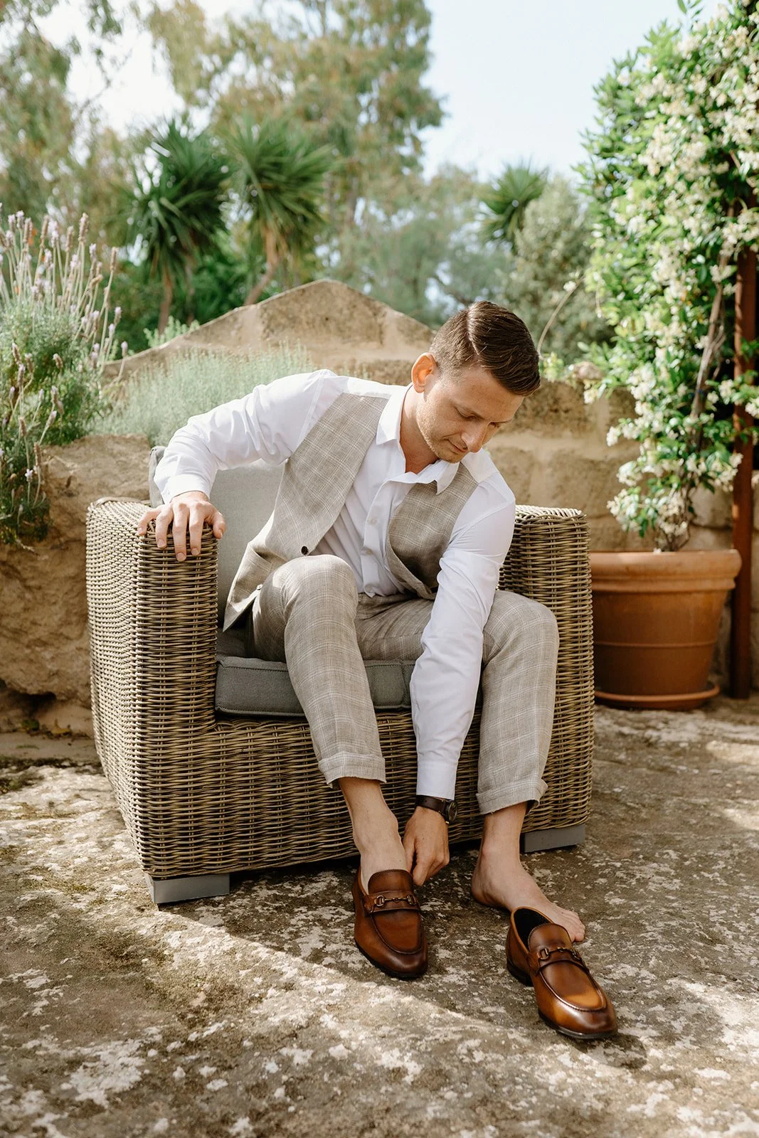 A man in a beige plaid suit and white shirt is sitting on a wicker chair outdoors, tying the laces of his brown loafers, surrounded by greenery and potted plants.