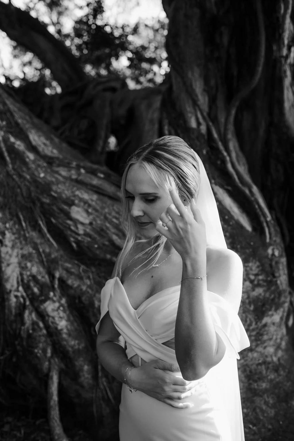 Black and white photo of a woman in a wedding dress standing outdoors near large tree roots. She is looking down with her hand near her face, with a contemplative expression.