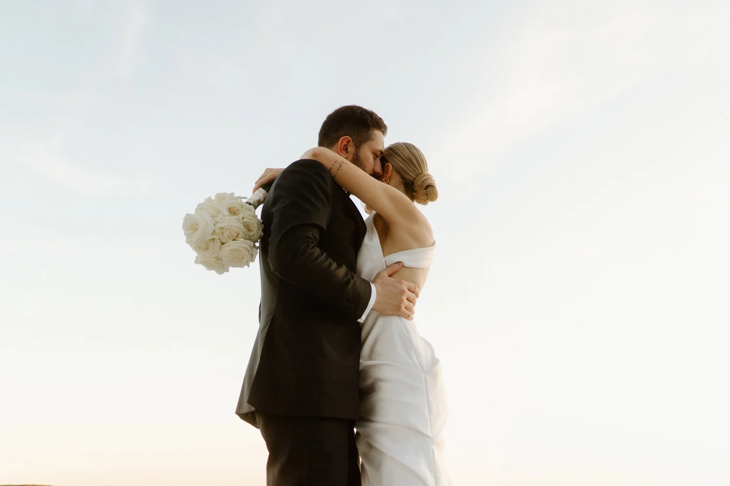 A couple dressed in wedding attire embracing and kissing outdoors, with the groom holding a bouquet of white roses behind the bride.