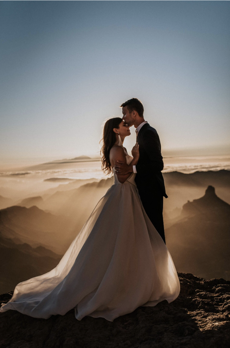 A bride and groom sharing a kiss on a mountain with a desert landscape in the background during sunset.
