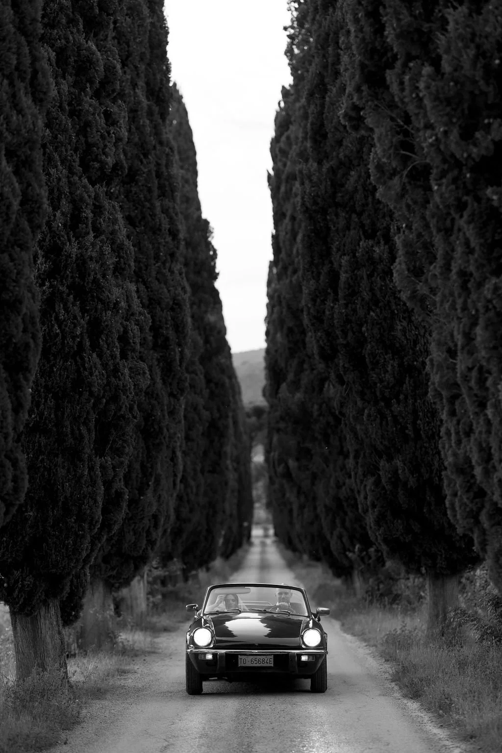 A black vintage sports car driving on a narrow dirt road lined with tall, dense trees on both sides.