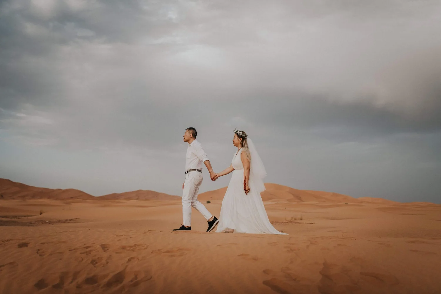 A couple holding hands walking in a desert landscape under a cloudy sky, with the woman in a white wedding dress and floral crown, and the man in a white shirt and light-colored pants.