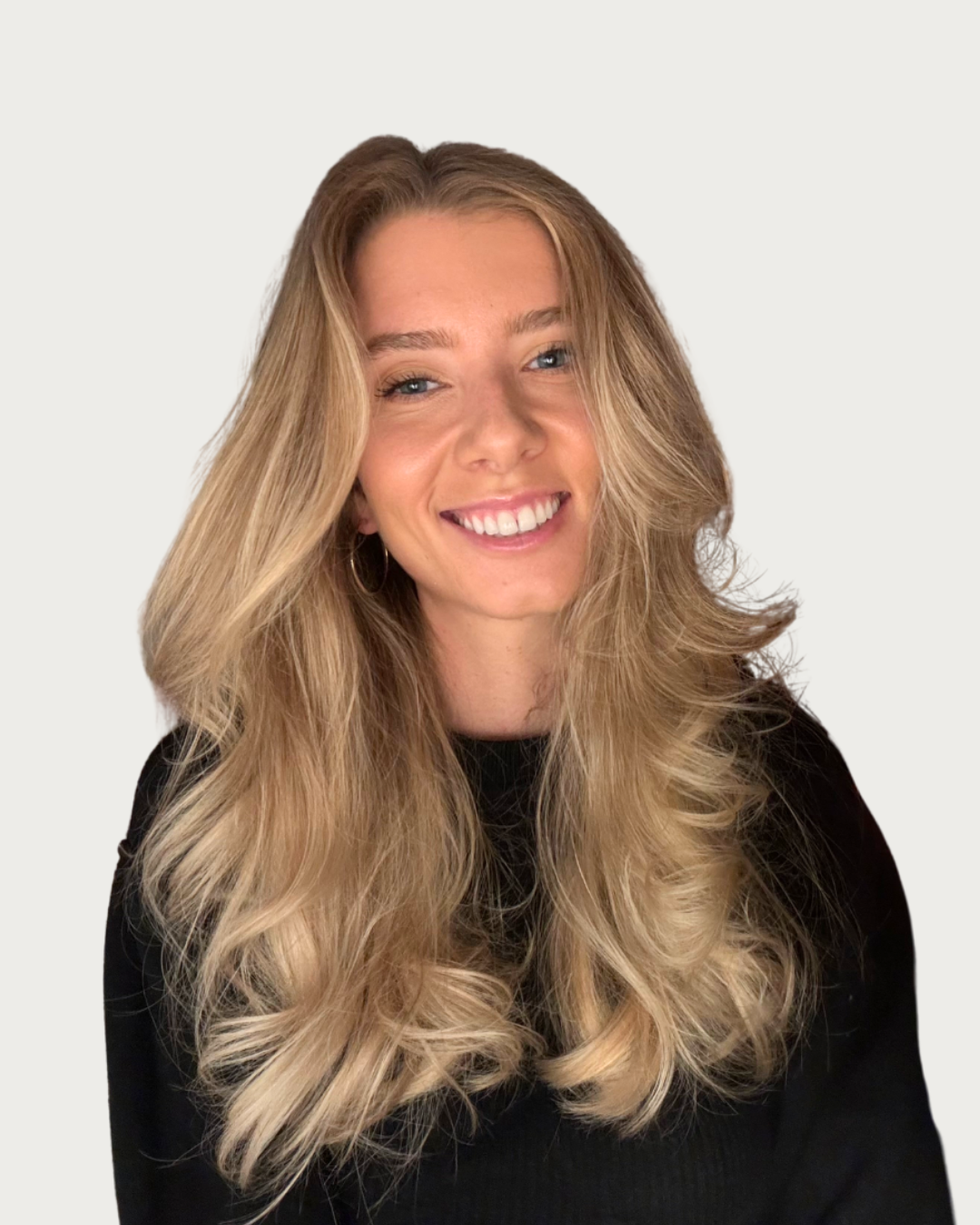 A woman with long, wavy blonde hair smiling, wearing a black top and hoop earrings, against a plain light background.