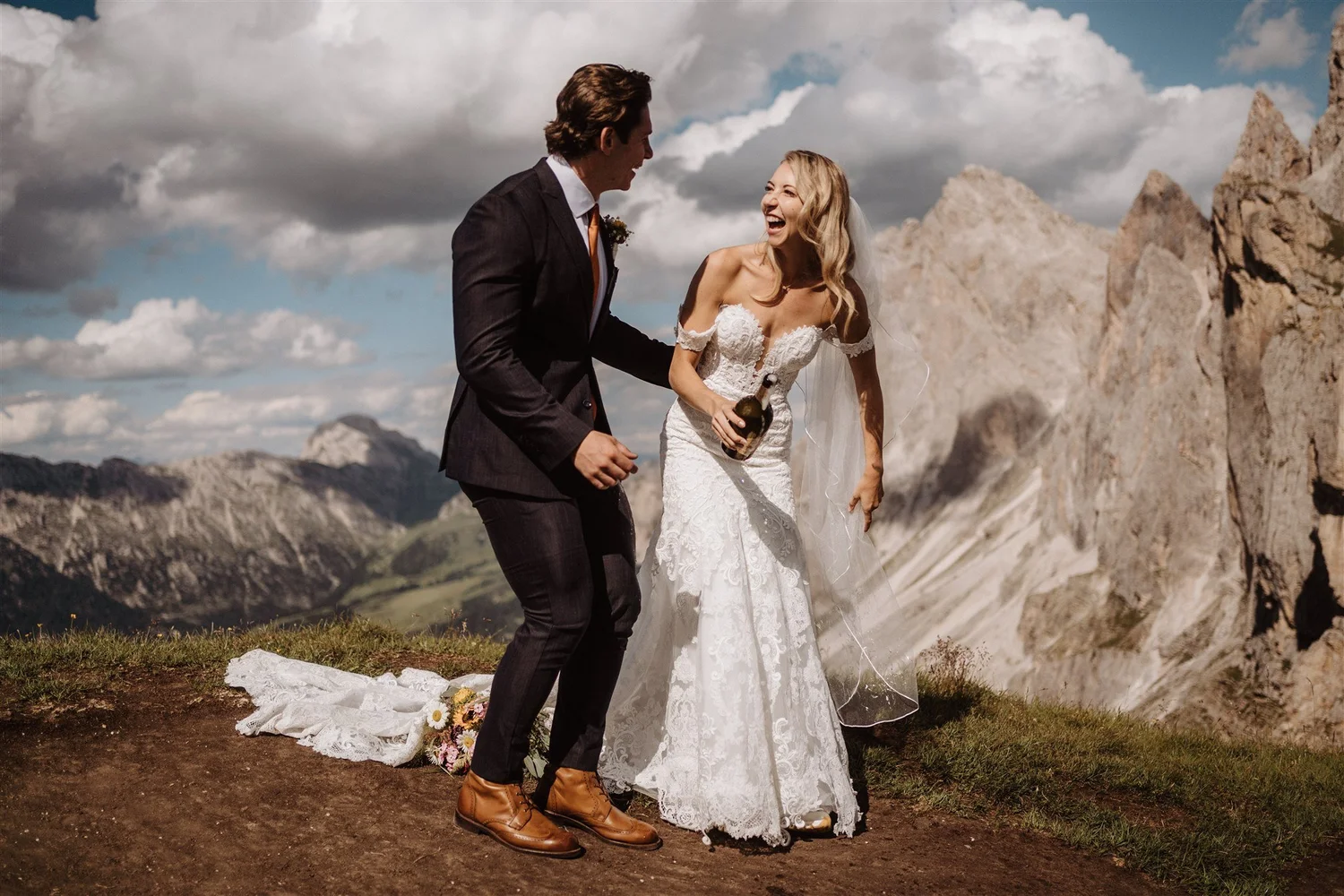 A bride and groom smiling and laughing outdoors, with mountains and a cloudy sky in the background. The bride is wearing a white lace wedding dress and holding a bottle, while the groom is dressed in a dark suit and brown boots.