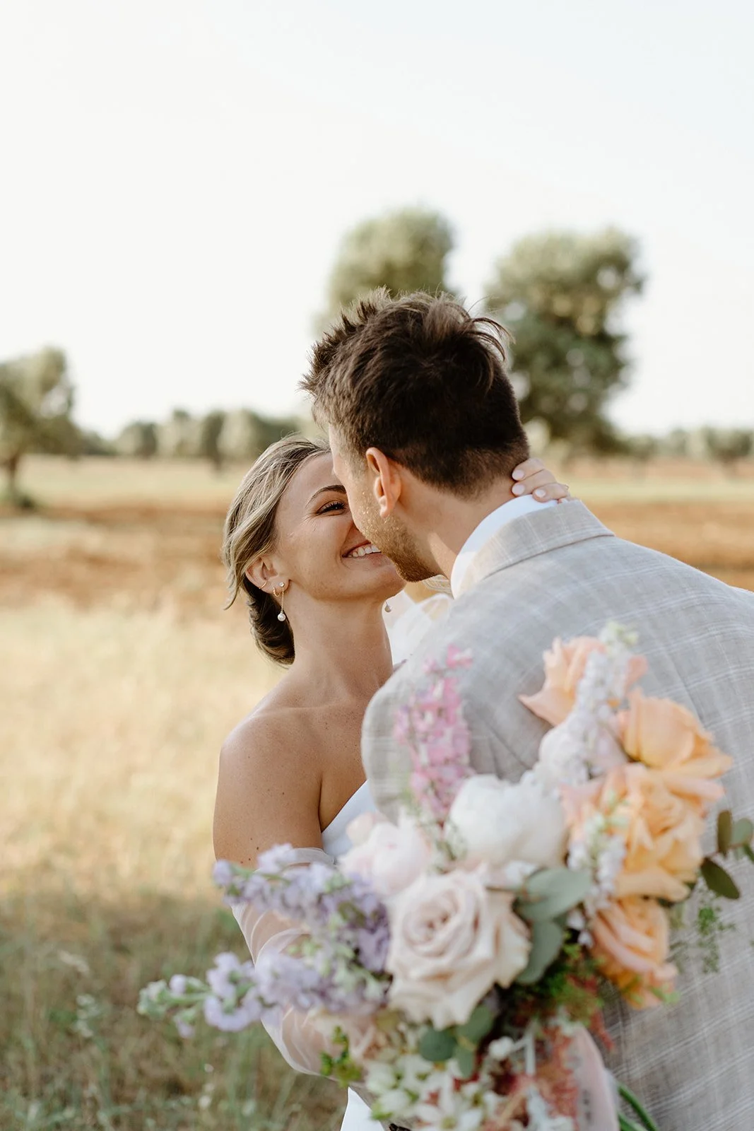 A bride and groom embracing outdoors, with their noses touching and smiling. The bride is holding a large bouquet of pastel-colored flowers, and the groom is wearing a light grey suit. The background features a field with trees under a bright sky.