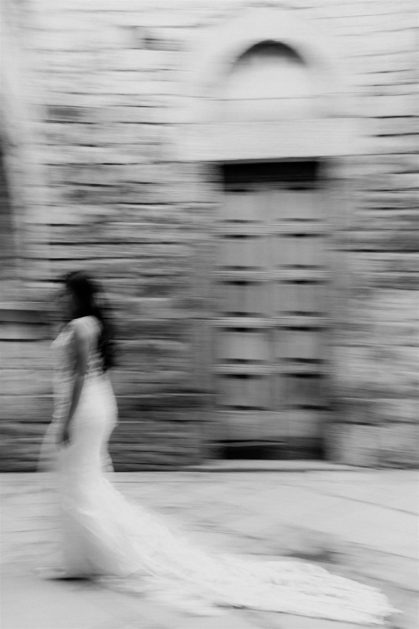 A woman in a long white dress walking on the street in front of a brick wall with an arched window. The image is blurred, creating a motion effect.