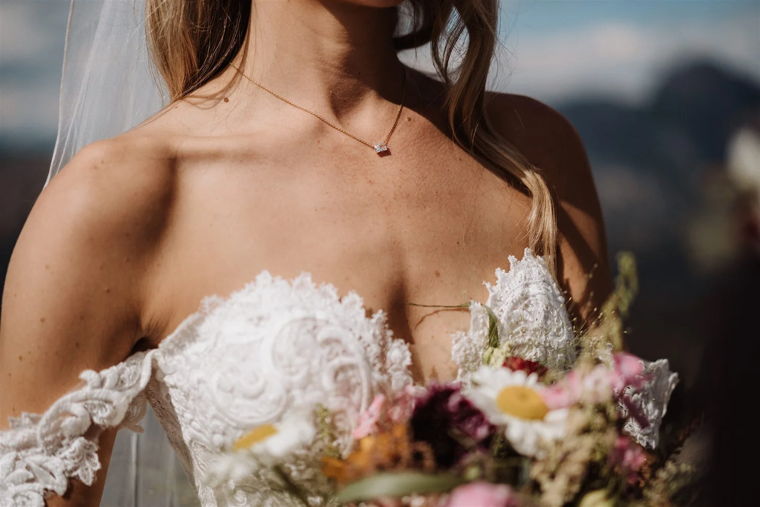 Close-up of a woman wearing a white lace off-the-shoulder dress and a gold necklace with a large gemstone. She has light brown hair and is holding a bouquet of colorful flowers, with a blurred background.