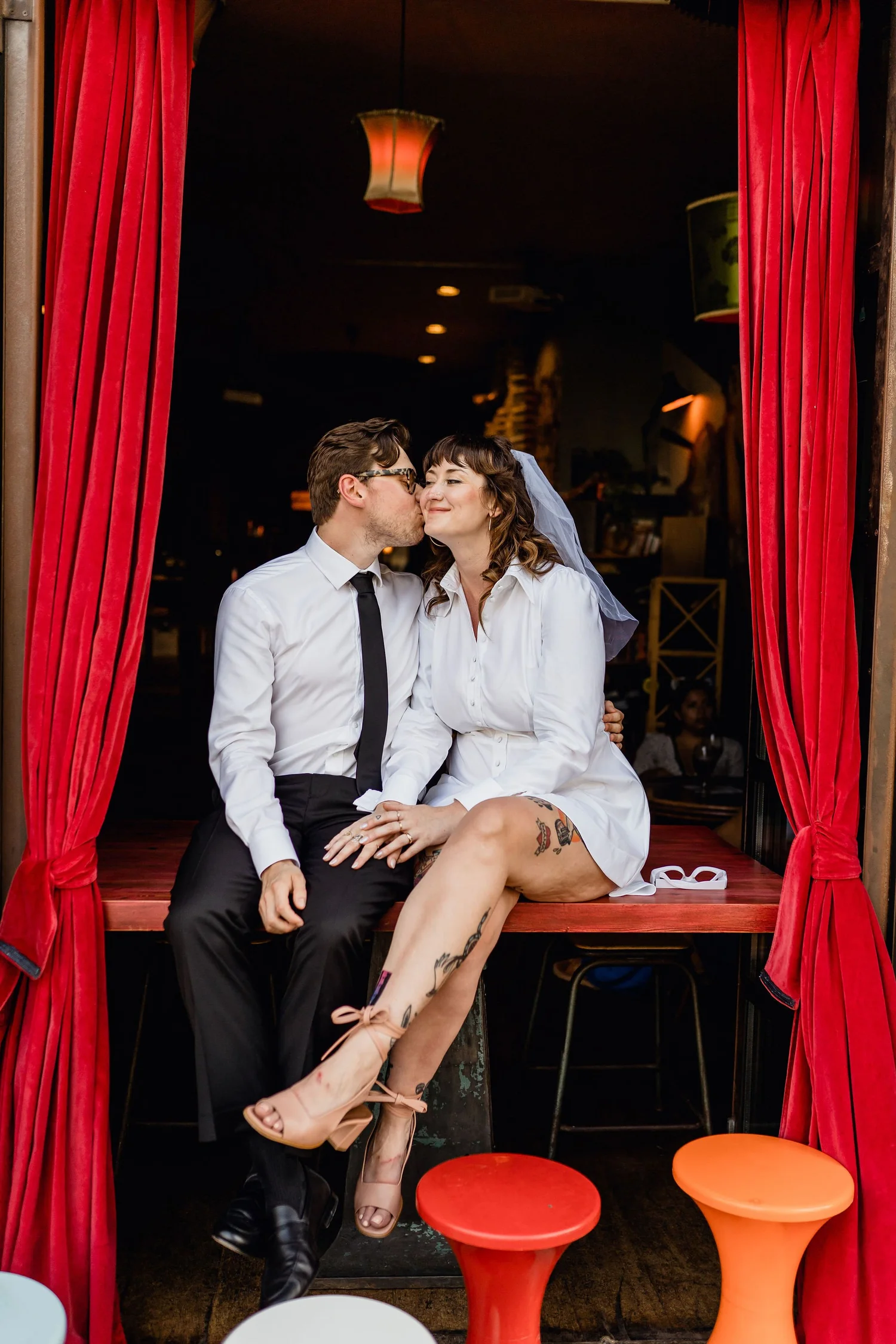 A couple dressed in wedding attire sitting on a stage with red curtains, kissing with their eyes closed. The woman has tattoos on her leg and is wearing heels and a veil, while the man is wearing glasses, a white shirt, and black trousers. There are