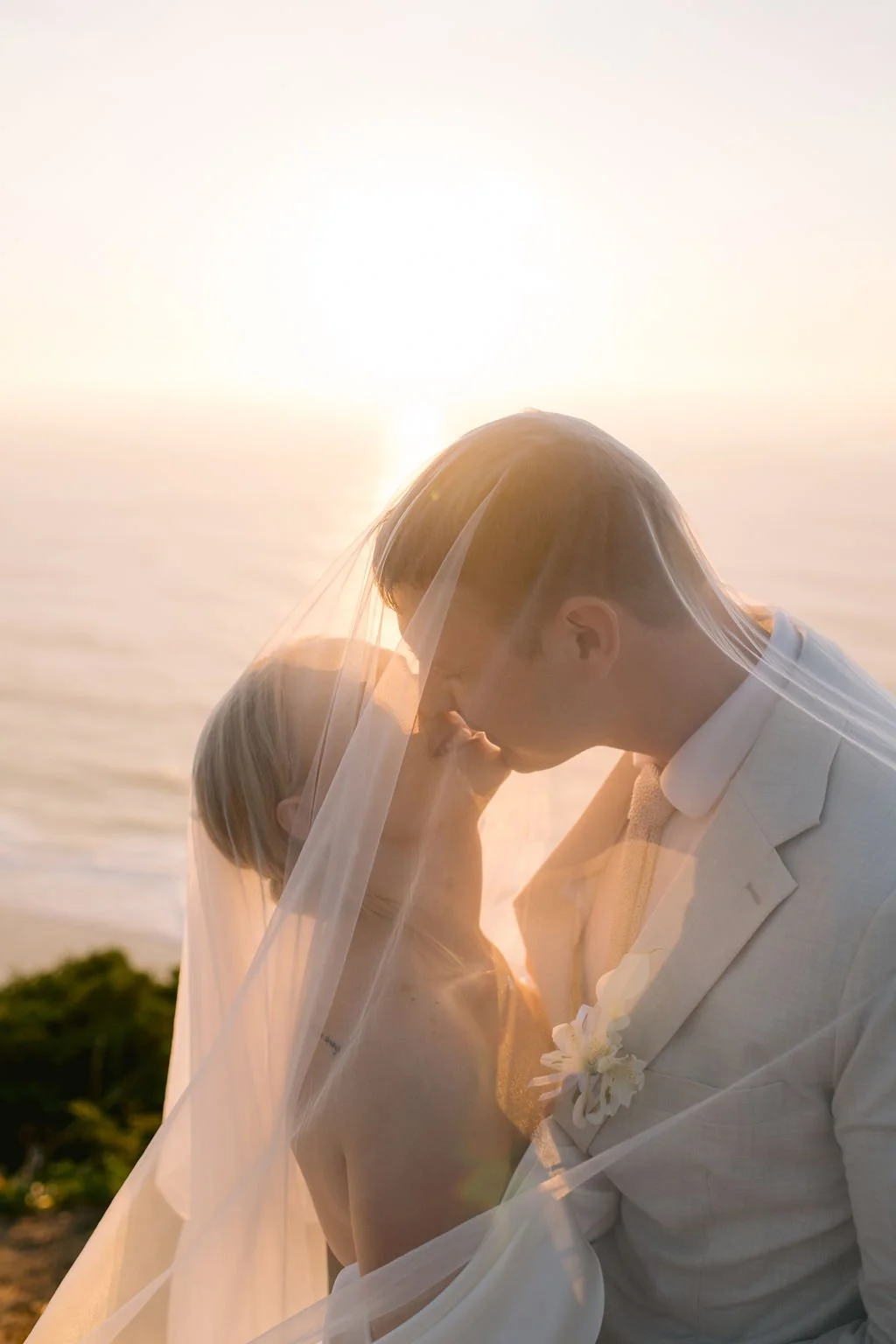 A couple dressed in wedding attire sharing an intimate moment under a veil with the sunset over the ocean in the background.