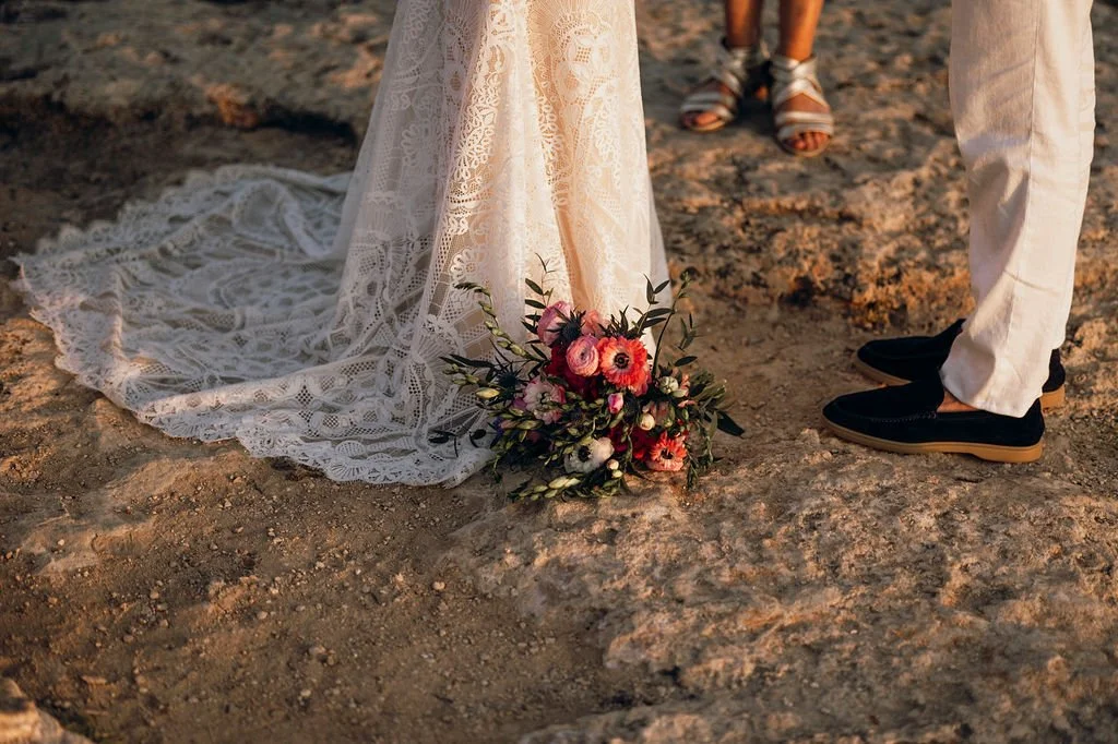Close-up of a bride's lace wedding dress train on uneven ground with a bouquet of pink and white flowers, and two people standing nearby with sandals and black shoes.