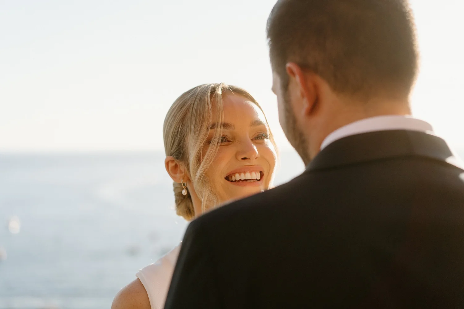 A woman and a man face each other outdoors near the ocean, smiling and laughing.