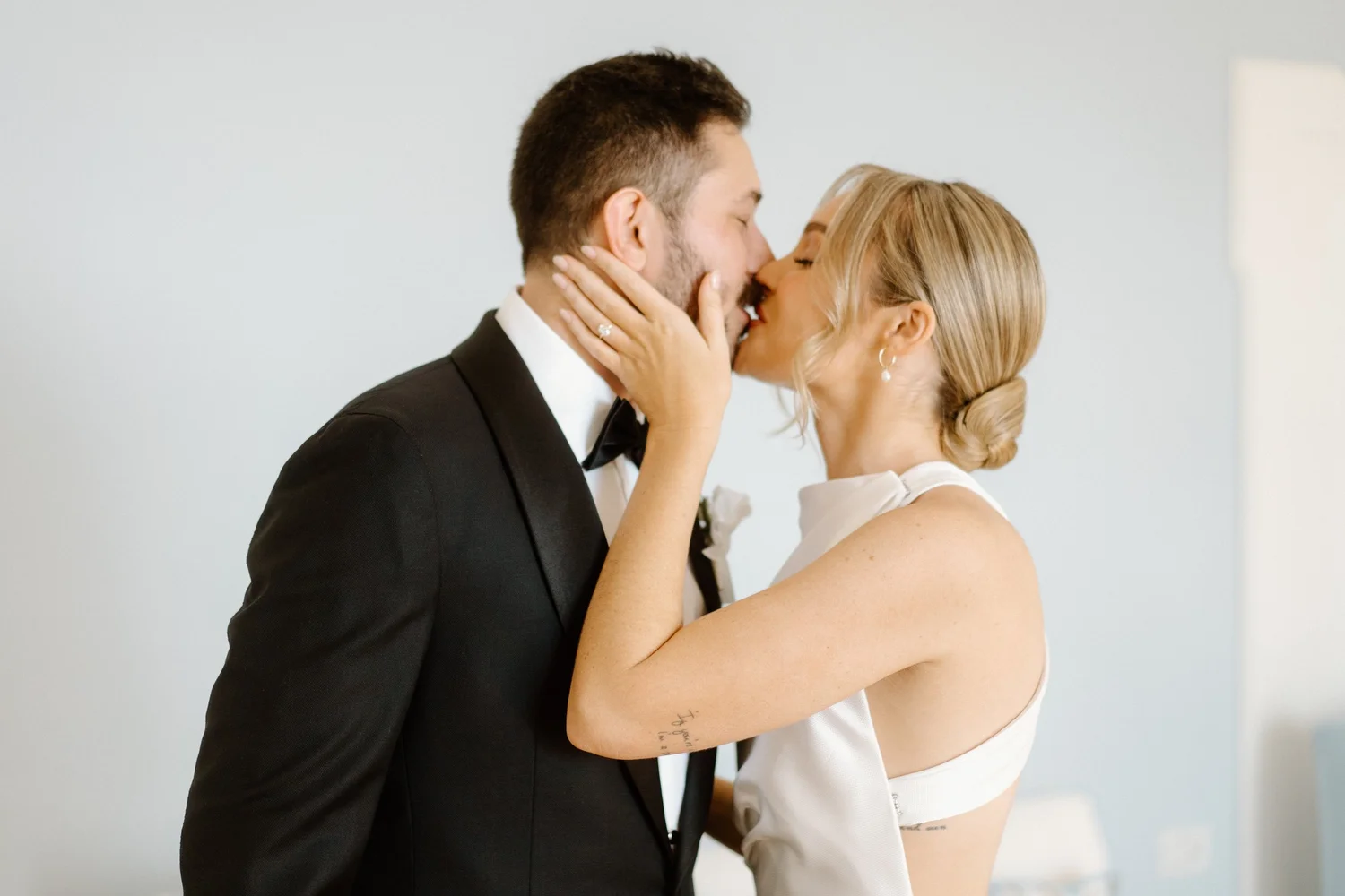 A couple kissing, the man in a tuxedo and the woman in a white dress, with their eyes closed in an intimate moment.