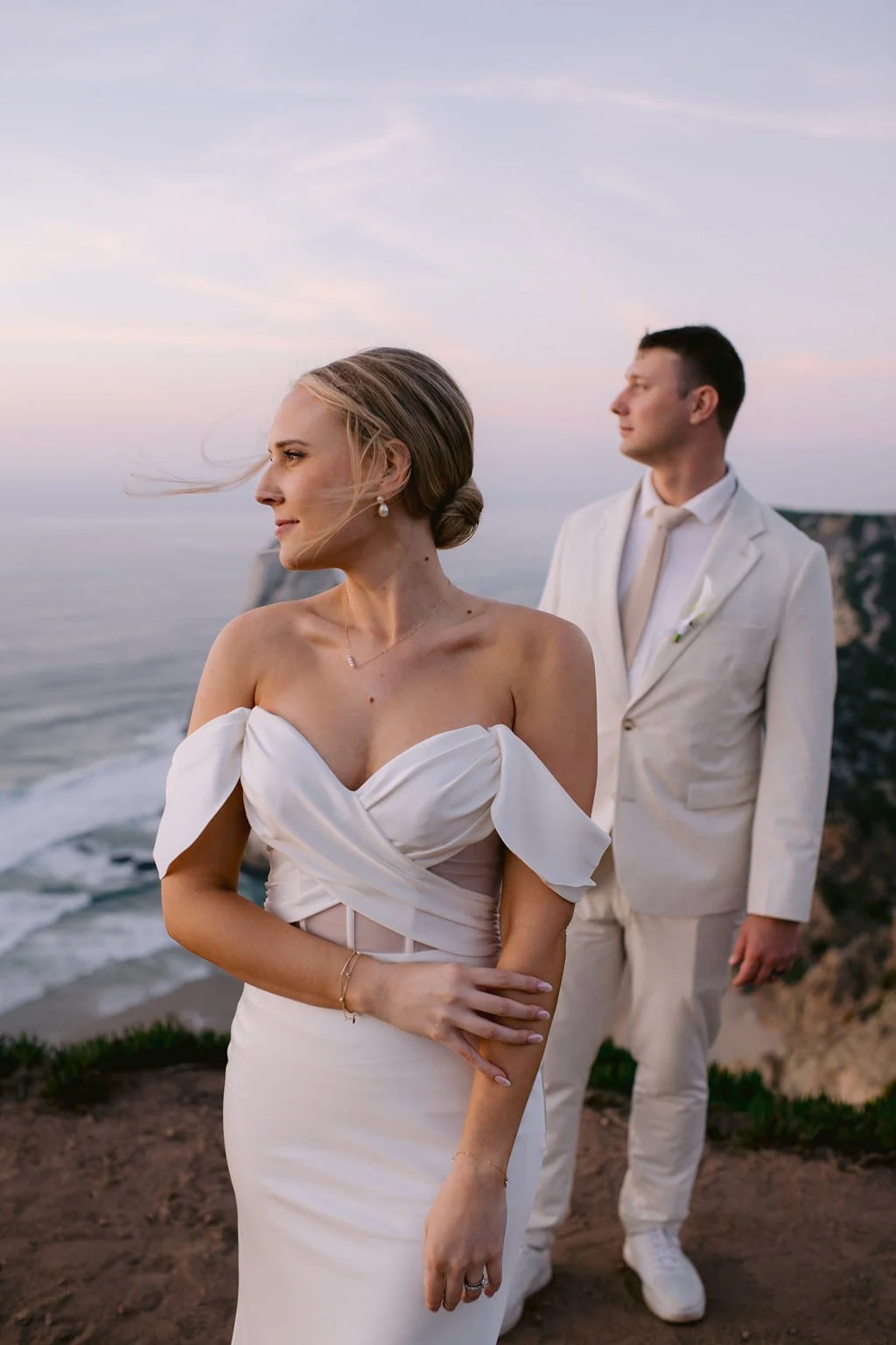 A bride in a white off-shoulder wedding dress and a groom in a white suit with a beige tie standing by the ocean at sunset.