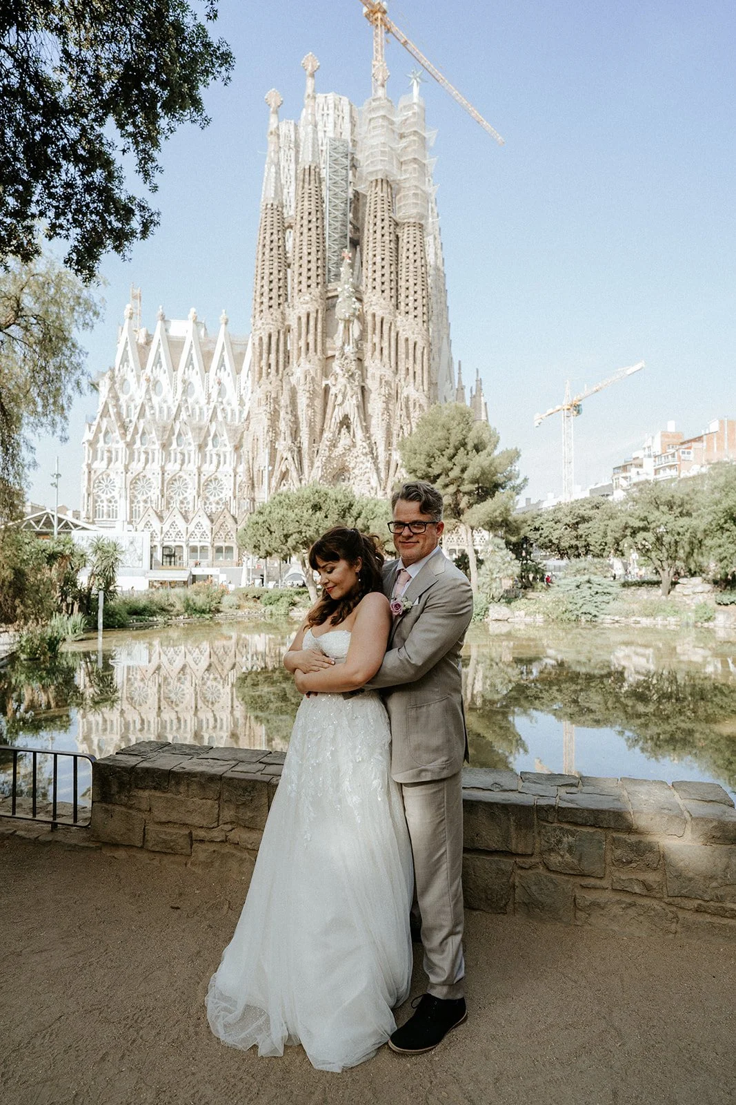 A couple in wedding attire standing in front of a pond with the Sagrada Familia cathedral under construction in Barcelona in the background, during daytime.