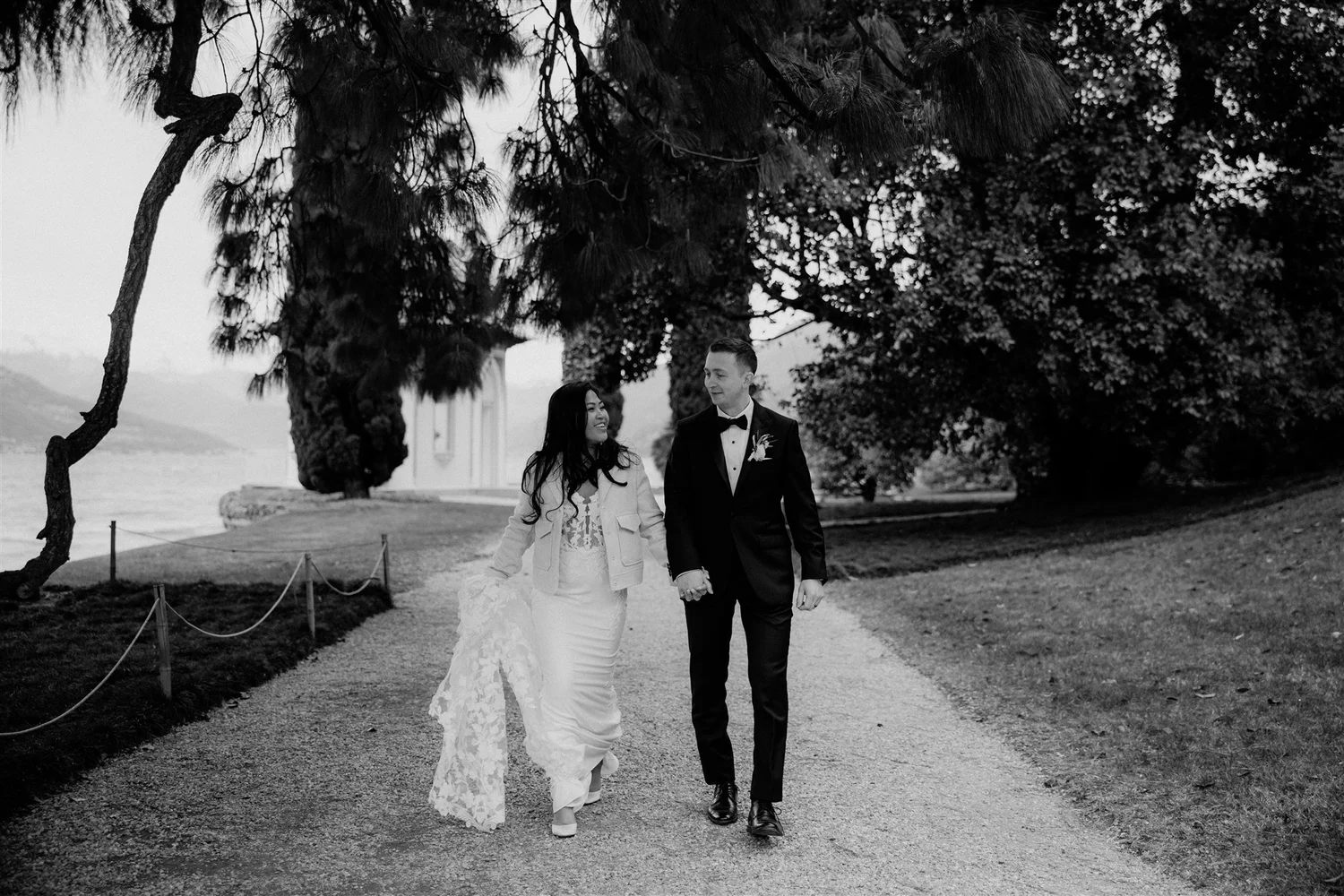 A black and white photo of a smiling bride and groom walking hand in hand outdoors under a large tree, dressed in wedding attire.