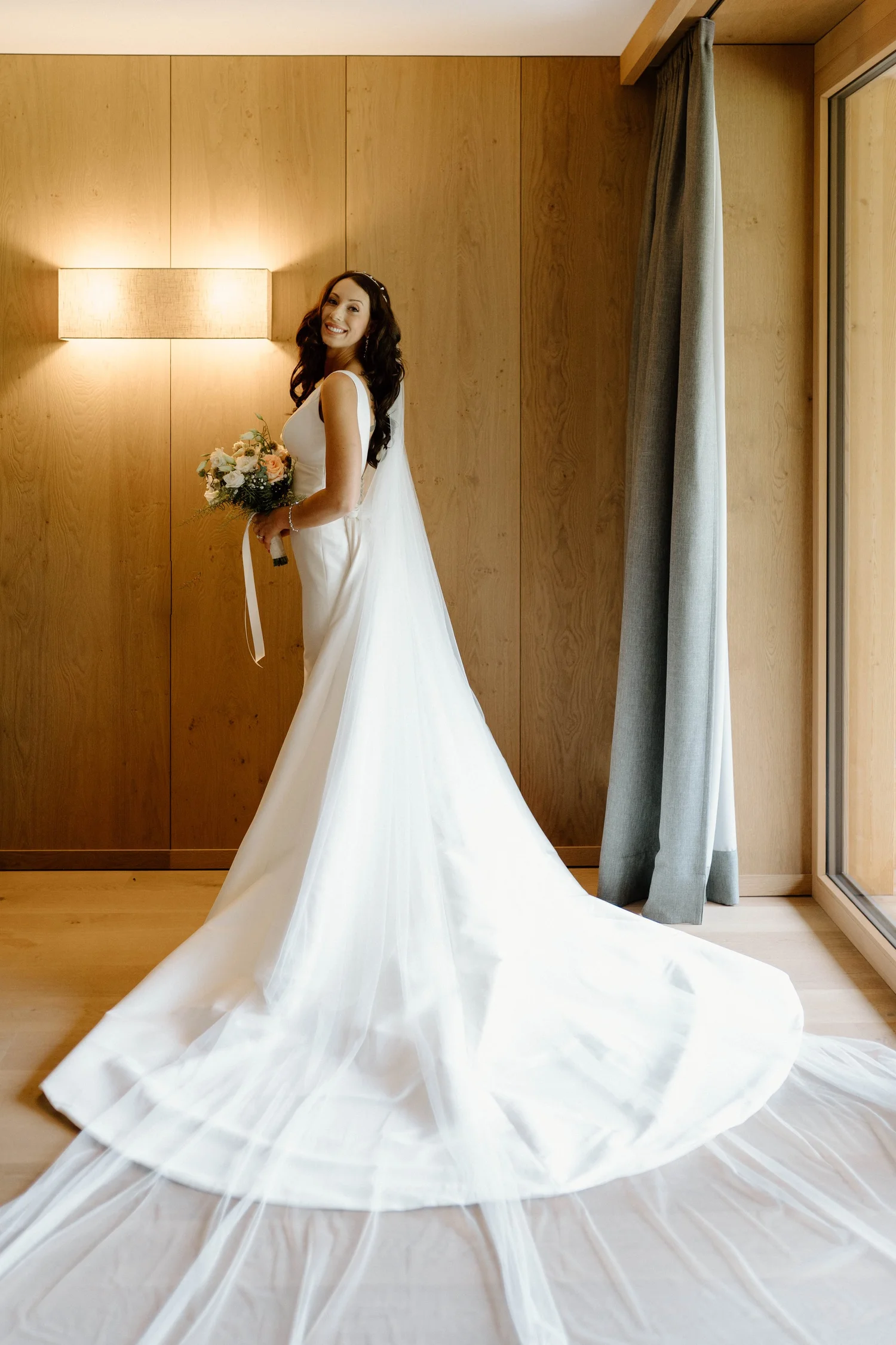 A woman in a white wedding dress holding a bouquet of flowers stands in a room with wooden walls and large window, smiling.