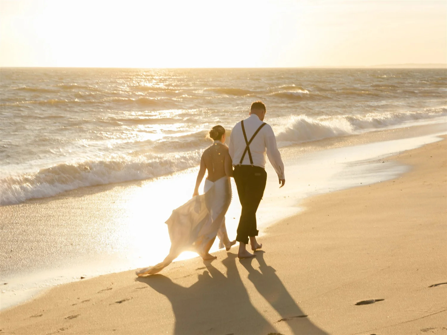 A couple dressed in wedding attire walking along the beach at sunset, with waves crashing nearby.