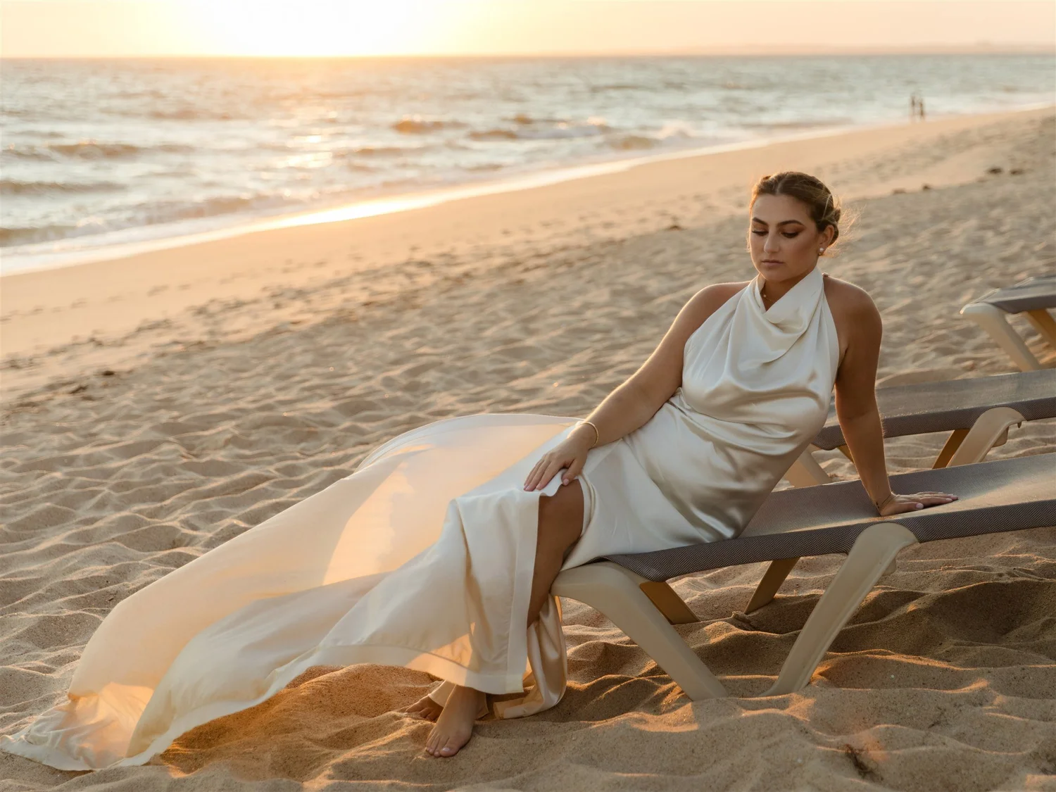 A woman in a white dress sitting on a lounge chair on the beach at sunset, looking downward with a calm expression.