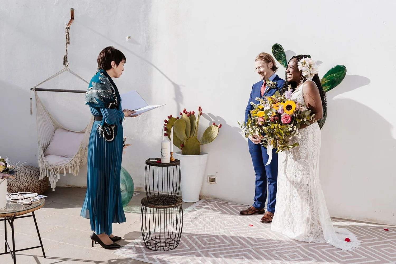 A couple getting married, holding a bouquet of flowers, standing in front of an officiant during a wedding ceremony on a sunny day.