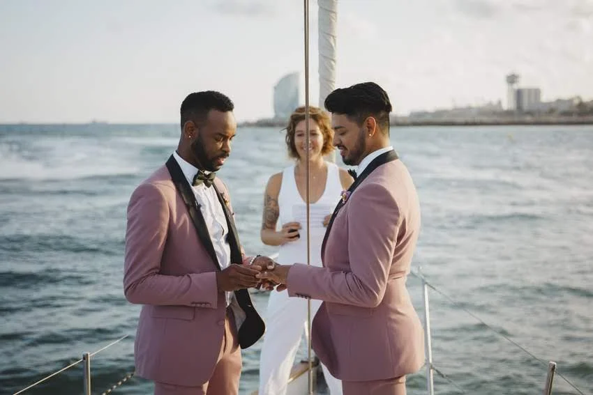 Two men in pink tuxedos exchanging rings on a boat, with a woman in a white dress holding a phone in the background, during a sunset by the ocean.