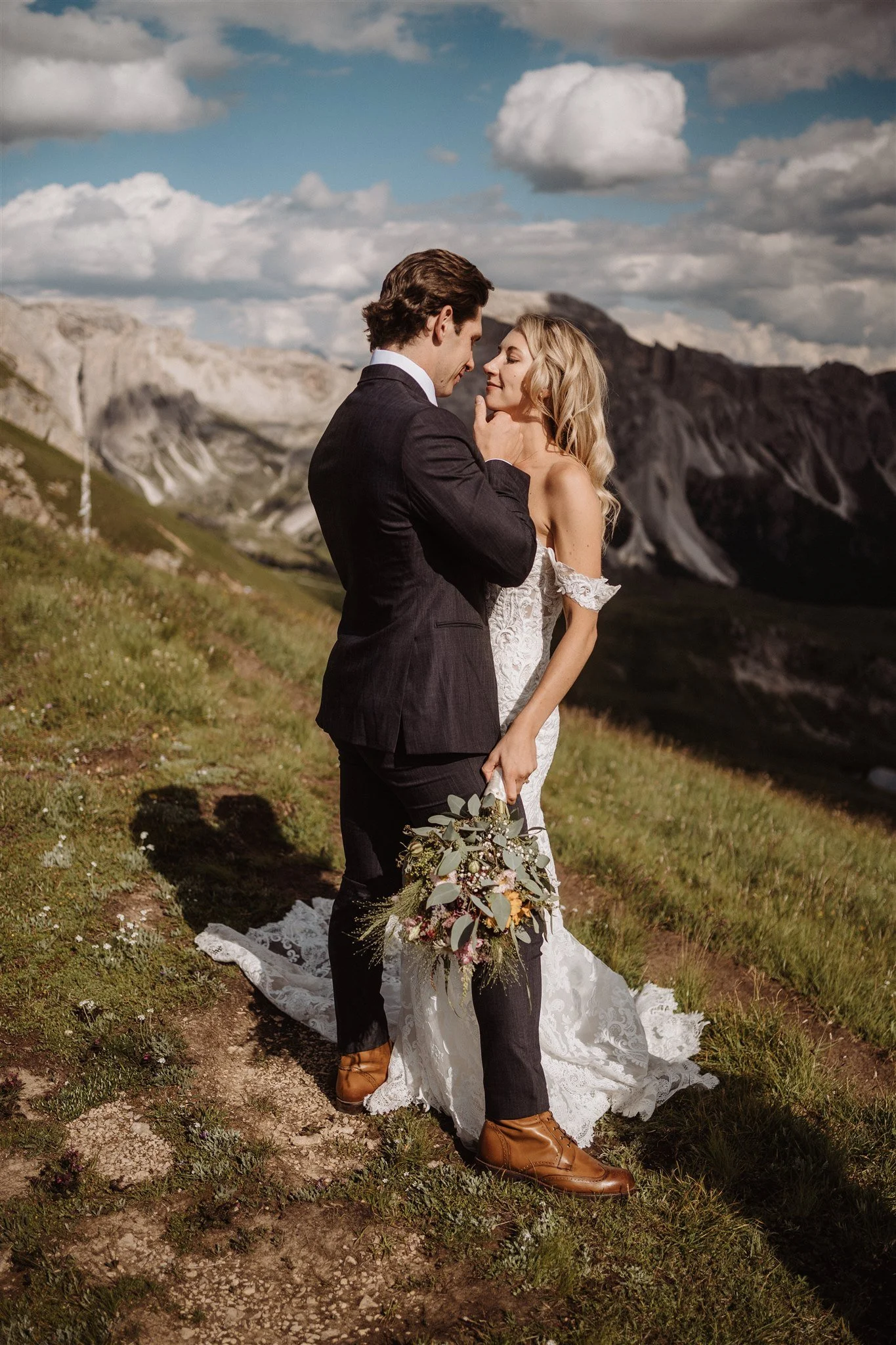 A bride and groom stand close together outdoors during daytime, with mountains and a cloudy sky in the background. The groom is touching the bride's chin, and they are looking into each other's eyes. The bride is wearing a lace wedding dress, holding