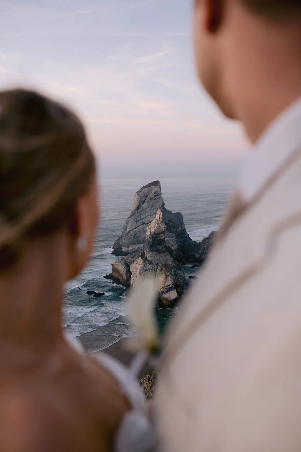 A couple on their wedding day looks at a large rock formation in the ocean during sunset.