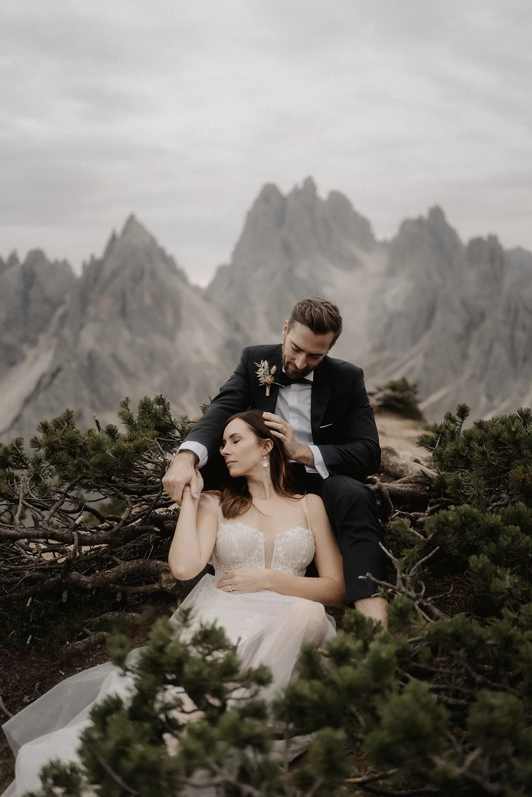 A couple dressed in wedding attire sitting outdoors among greenery, with mountains in the background, featuring a woman in a white gown and a man in a black suit.