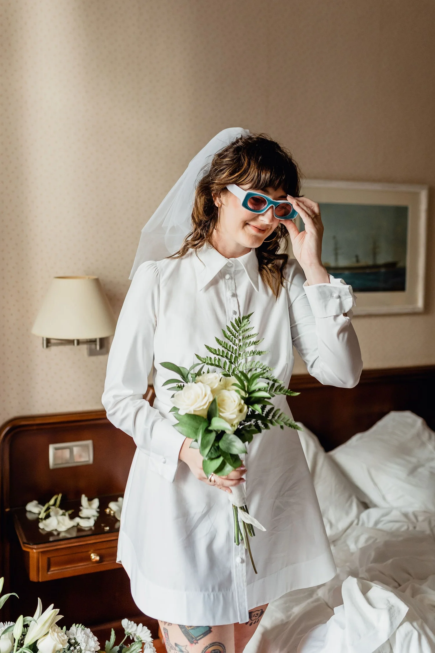 Woman in a white dress and veil holding a bouquet of white roses with greenery, standing in a bedroom with a nightstand, lamp, and picture frame.