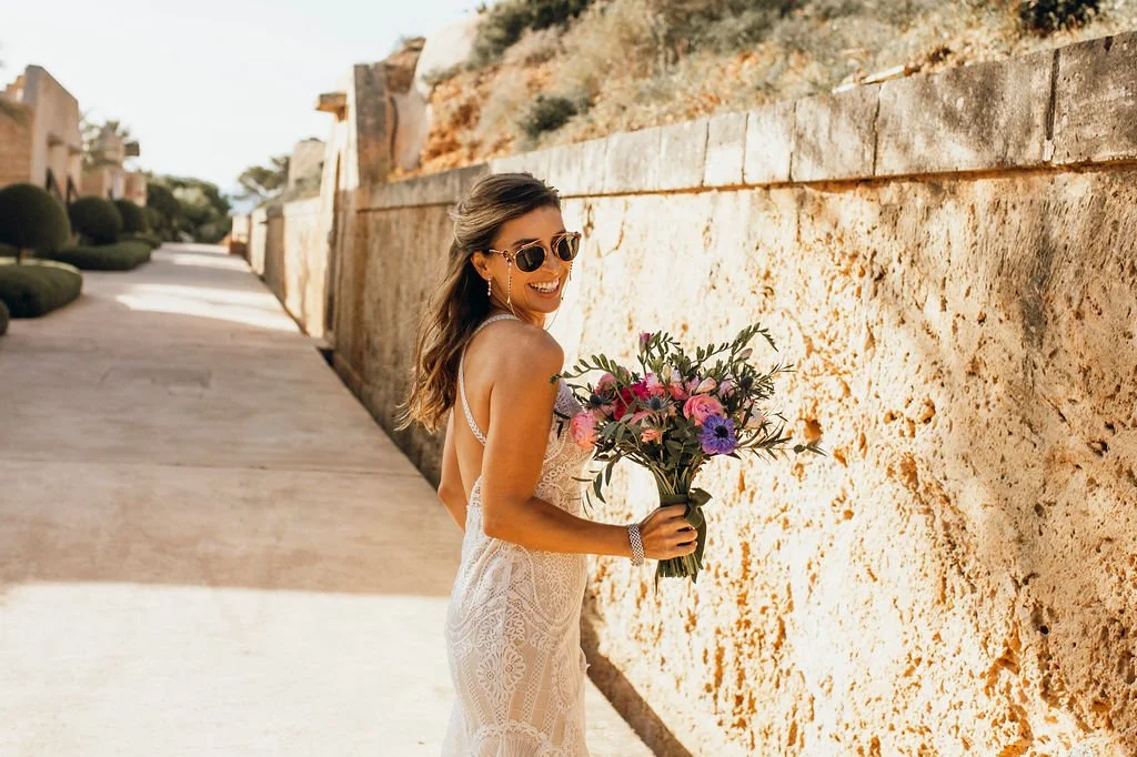 A woman in a white lace dress holding a bouquet of pink, purple, and green flowers, standing next to a beige stone wall outdoors, smiling and wearing sunglasses.