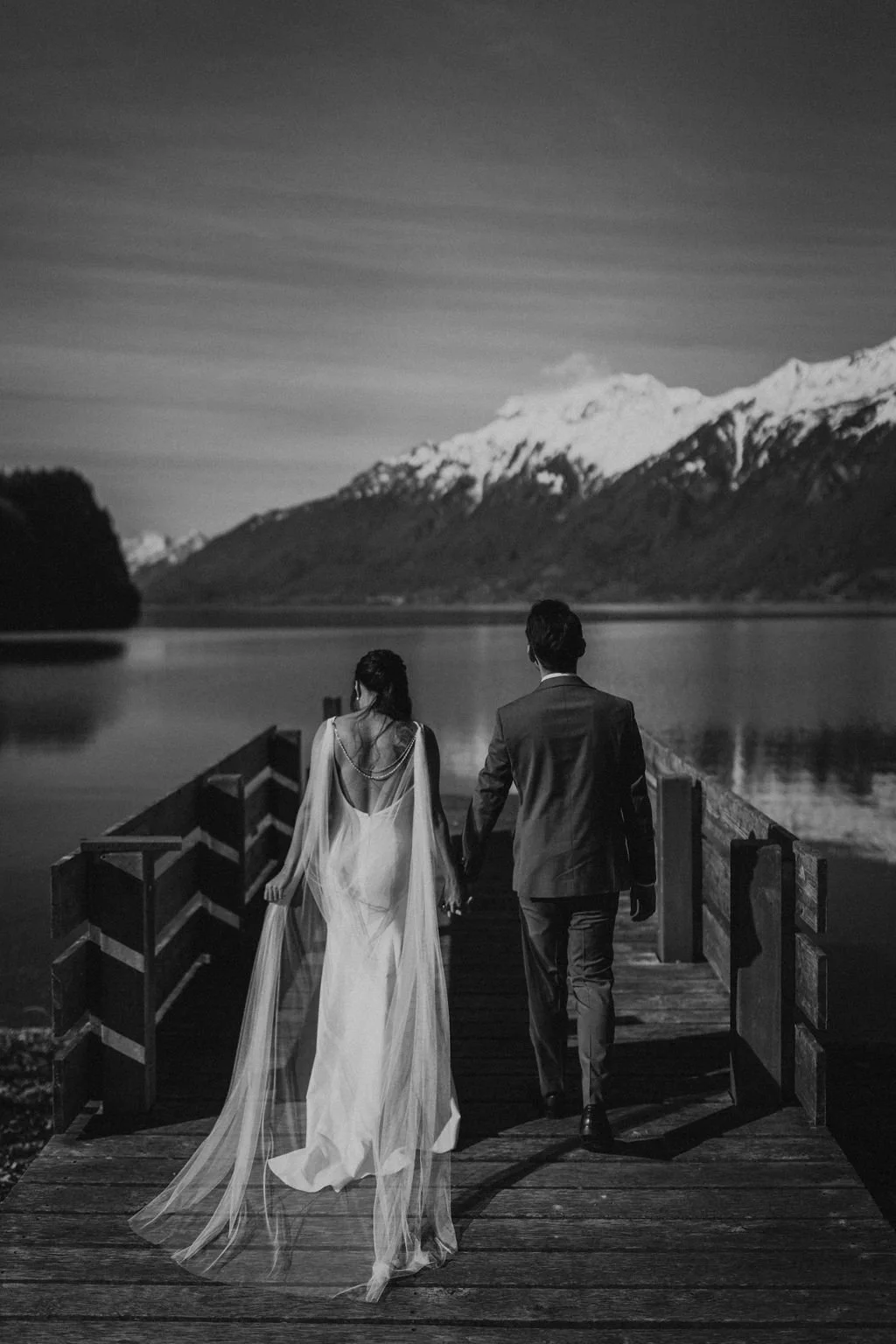 A black and white photo of a couple walking on a wooden dock by a lake with snow-capped mountains in the background. The woman is wearing a flowing wedding dress, and the man is dressed in a suit.