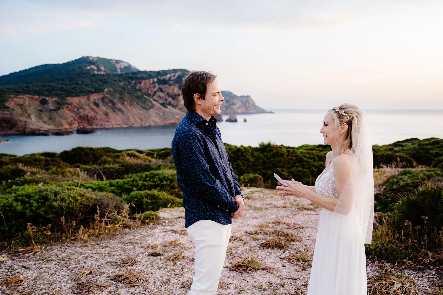 A couple getting married outdoors near the coast, with a man in a patterned blue shirt and white pants and a woman in a white bridal gown with a veil and hair accessory, standing on a rocky terrain with greenery, ocean, and cliffs in the background.
