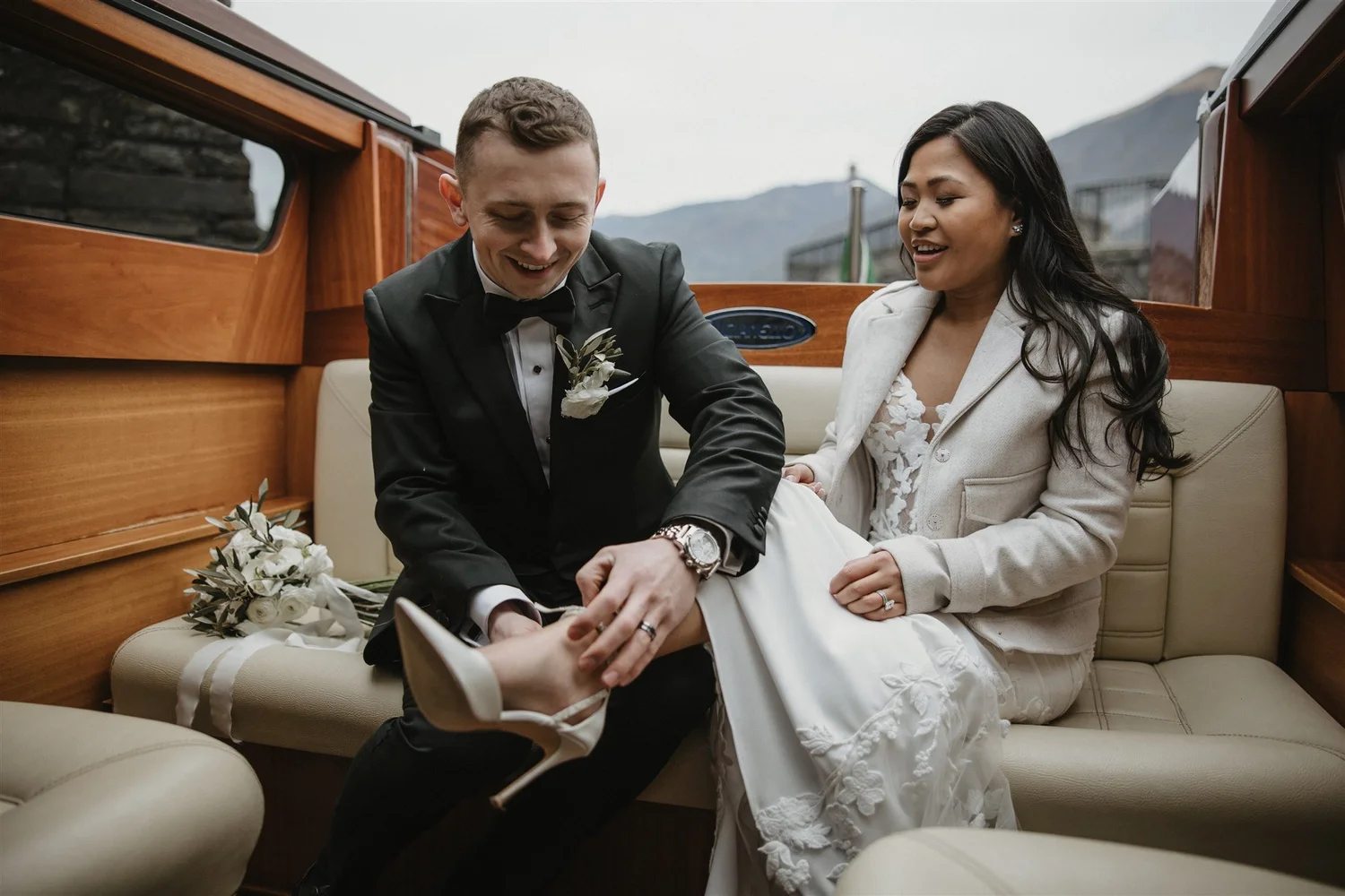 A newlywed couple sitting on a cream-colored cushioned sofa on a boat, smiling and looking at each other. The groom is wearing a black tuxedo with a bow tie and a white boutonniere. The bride is wearing a white wedding dress with a matching jacket, a